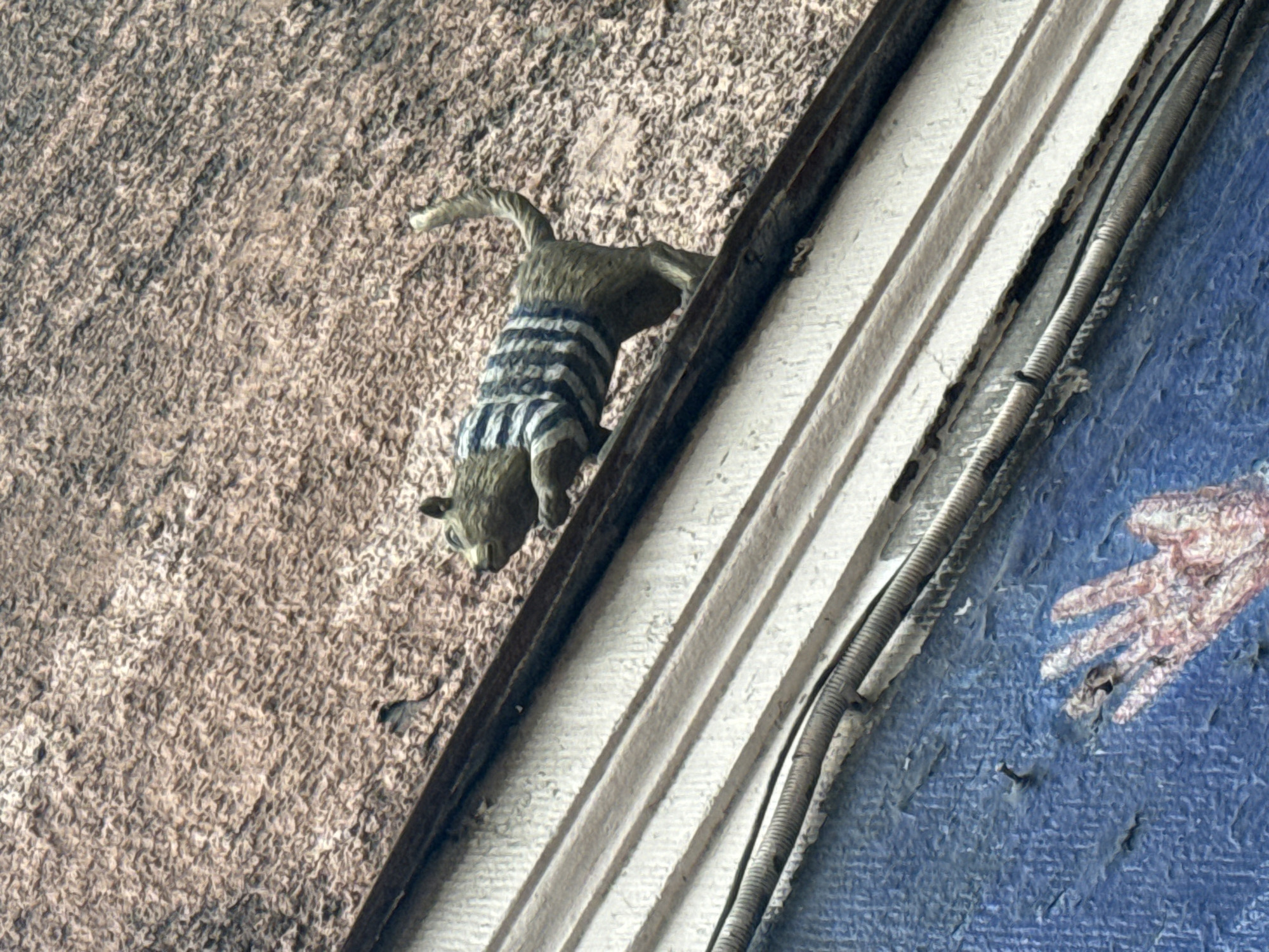 A dilapidated exterior corner is visible in the image. A sculptural representation of a squirrel in a striped sweater is affixed to a rough, beige-colored wall. Adjacent to the squirrel is a white, tiered architectural molding. To the right of the molding, a blue-painted surface displays a faded depiction of a human hand. Electrical conduits run along the edge of the molding. The location is Saint Petersburg, Russia.