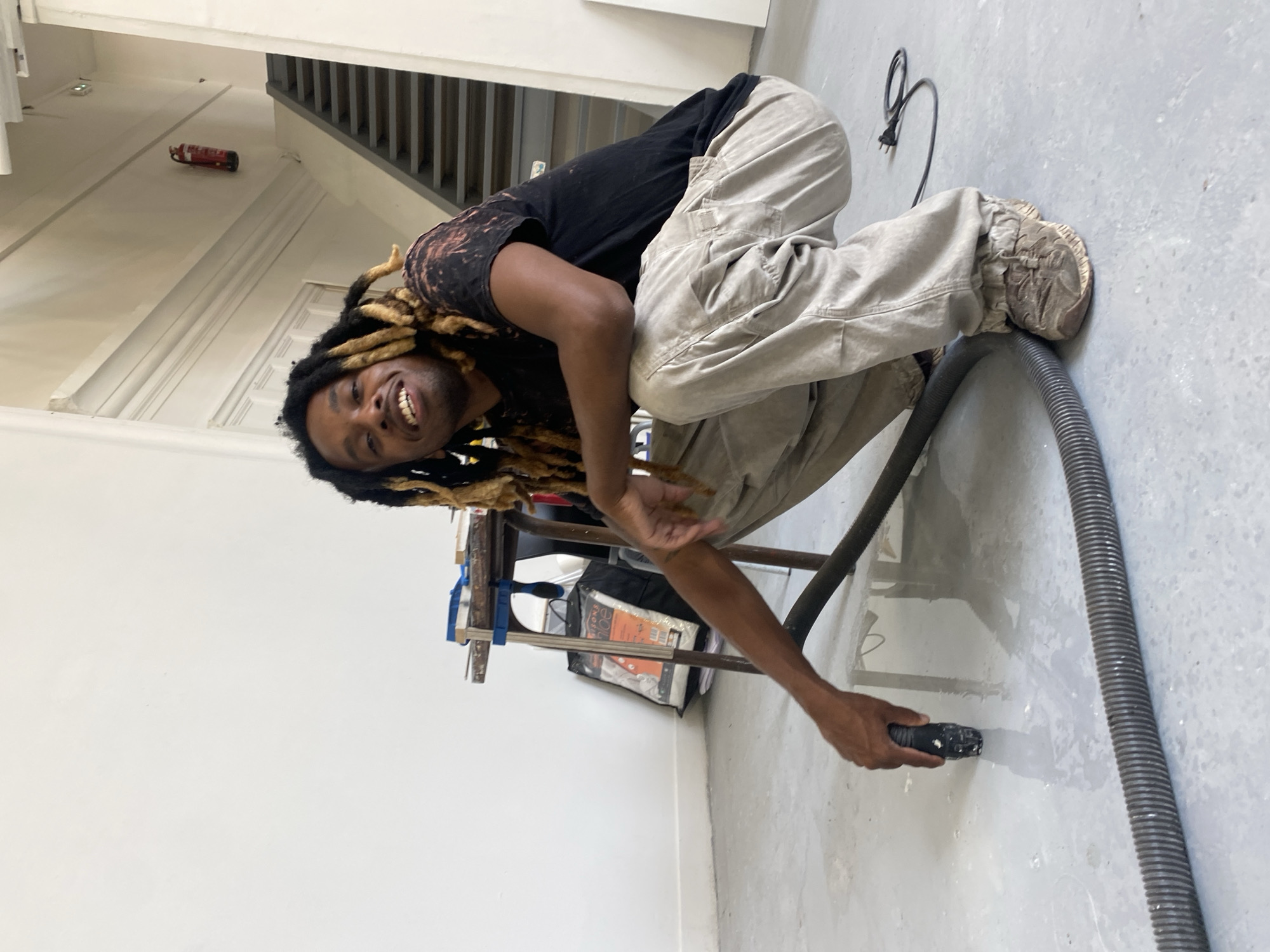 The image shows a Black man with dreadlocks, wearing a black t-shirt and tan work pants, crouching on a concrete floor. He is smiling at the camera and holding a vacuum cleaner hose with a nozzle in his right hand, directing it towards the floor. A black electrical cord with a plug is visible on the floor behind him.
In the background, there is a metal staircase with gray railings and a white wall. A red fire extinguisher is mounted on the white wall. To the left of the man, there is a workbench with clamps and what appear to be art supplies or tools. Bags and papers are stacked on the floor near the workbench. The scene appears to be indoors, possibly in a studio or workshop. The lighting suggests it is daytime. The overall mood seems relaxed and positive due to the subject's smile. No visible text is present in the image. The provided location context is Paris, France, but this information cannot be visually confirmed from the image itself.