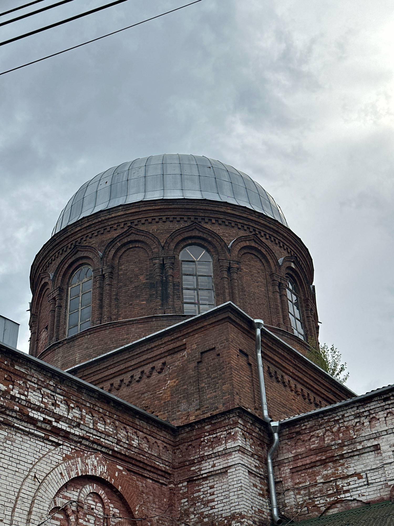 This is a low-angle, medium shot of a large, domed brick building under an overcast sky. The building appears to be an old church or similar structure, possibly with religious significance. The dome is made of metal panels, weathered and slightly discolored, and it sits atop a circular brick base. The brickwork of the base is a deep red-brown and shows signs of age, with some areas of wear and discoloration, particularly on the lower section of the building which is partially whitewashed.
The dome features a series of arched windows with multiple panes, framed by decorative brickwork. These windows are tall and narrow, with the glass appearing somewhat opaque. Below the dome, the building has a stepped architectural design, also constructed from brick. A metallic downspout runs vertically along one of the corners of the building. The sky above is a uniform pale gray, with some indistinct cloud formations and the faint glint of sunlight breaking through. Several thin, dark wires are visible against the sky in the upper left portion of the image.
The scene is devoid of people and any apparent activity. The overall impression is one of aged architecture under a somber sky. The style of the building, particularly the dome and arched windows, is characteristic of Orthodox Christian churches found in Russia. This specific building is identified as the Church of the Resurrection of Christ in Biysk, Russia.