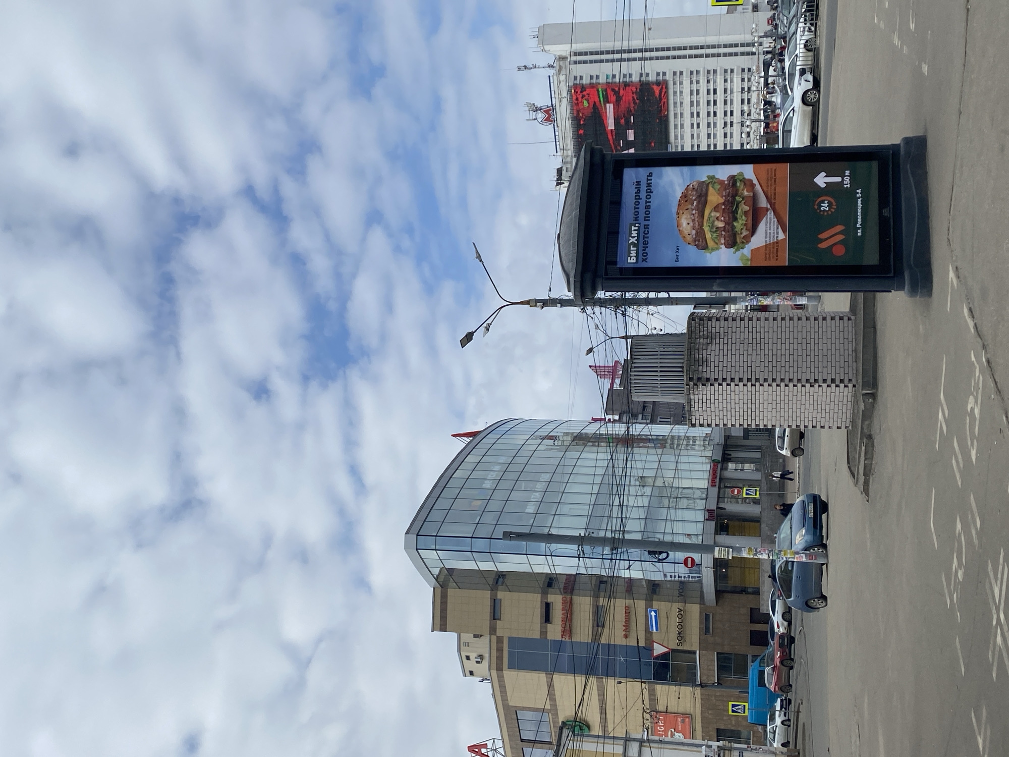An urban street scene in Nizhny Novgorod, Russia, is captured on what appears to be a cloudy or partly cloudy day. The image is oriented vertically but depicts a horizontal panorama of the street and buildings.
In the foreground, a prominent digital billboard displays an advertisement for a burger called "Биг Хит" (Big Hit) with the tagline "который хочется повторить" (which you want to repeat). The ad also indicates "24ч" (24 hours), an address "пл. Революции, 5-А" (Revolution Square, 5-A), and a distance of "150 м" (150 m), along with a fast-food chain logo (resembling the Vkusno i Tochka brand that replaced McDonald's in Russia). To the left of the billboard, a street light pole with multiple lamps extends over the street.
The midground features a modern building with a curved, reflective glass facade on the left. Below it, other commercial buildings are visible with signs such as "Monro," "SOKOLOV," "Vdox," and partially visible "LIGH" (likely "LIGHT"). Several cars are parked along the street, and a few pedestrians can be seen walking. Power lines crisscross the frame, adding to the urban feel.
In the background, taller, more traditional-looking buildings rise, with one featuring a large digital screen displaying red and black abstract graphics. The sky above is mostly covered with scattered white clouds against a blue backdrop. The overall impression is that of a bustling city center, characteristic of a Russian metropolitan area.