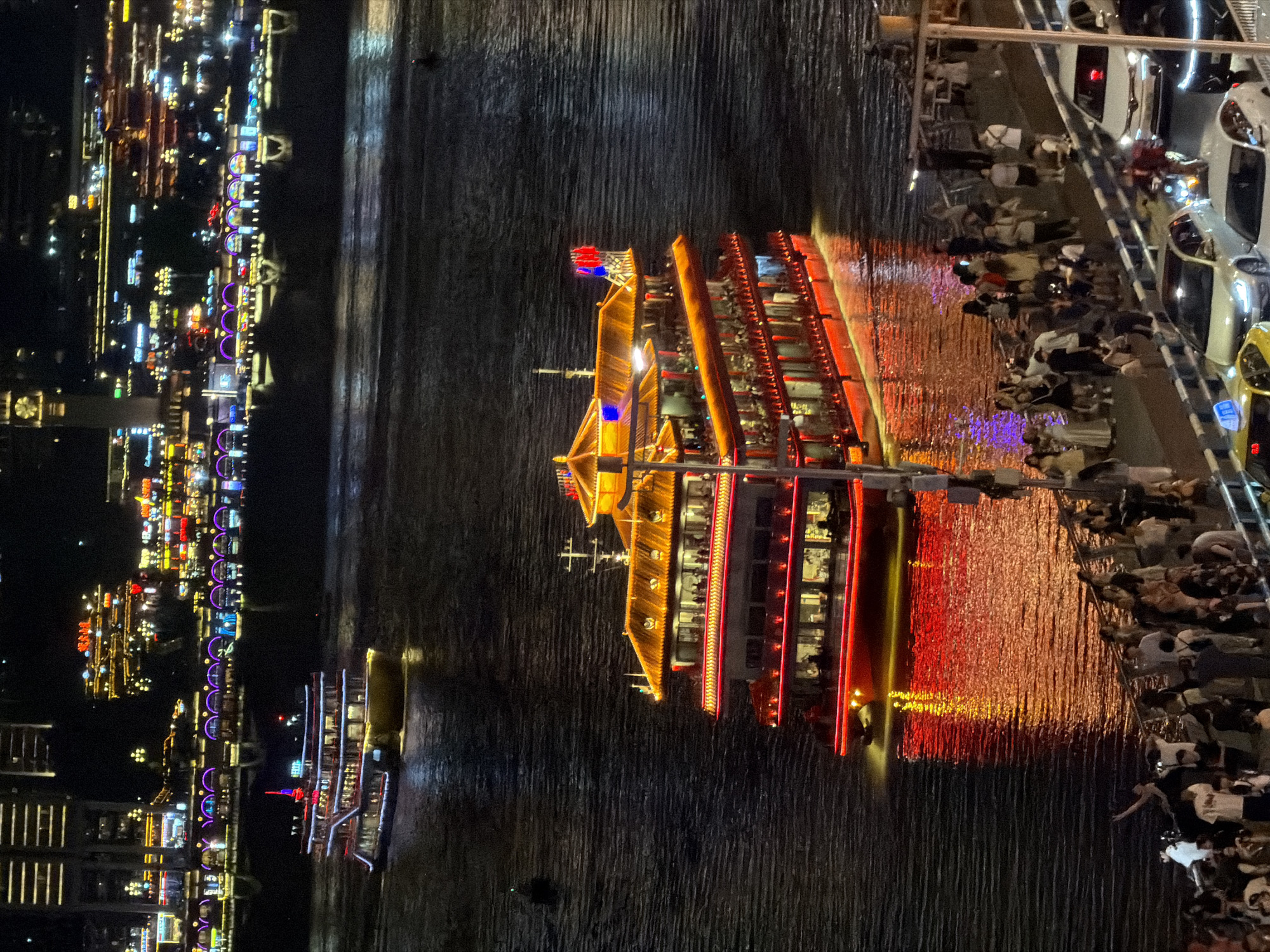 The image captures a nighttime scene along the Yangtze River in the Yuzhong District of Chongqing, China. The foreground shows a bustling riverside promenade crowded with pedestrians, many of whom are casually dressed and appear to be observing the river and the illuminated buildings across the water. Several cars are parked or driving on the road adjacent to the promenade, including a white sedan and a yellow taxi.
In the middle ground, a large, multi-decked river cruise ship is prominently displayed, adorned with bright red and yellow lights. The ship has a traditional Chinese architectural style with tiled roofs. Numerous people are visible on the decks of the ship, suggesting it is either embarking, disembarking, or underway with passengers. The ship's red hull is reflected in the dark, rippling water, creating a vibrant streak of light.
In the background, the Chongqing skyline is illuminated by a multitude of colorful lights from various buildings and structures. The cityscape is dense and appears to rise steeply from the riverbank. Another, smaller yellow and white boat is seen further upriver, also illuminated, and moving away from the viewer. The overall impression is one of a lively urban environment at night, with significant river traffic and a vibrant city backdrop.