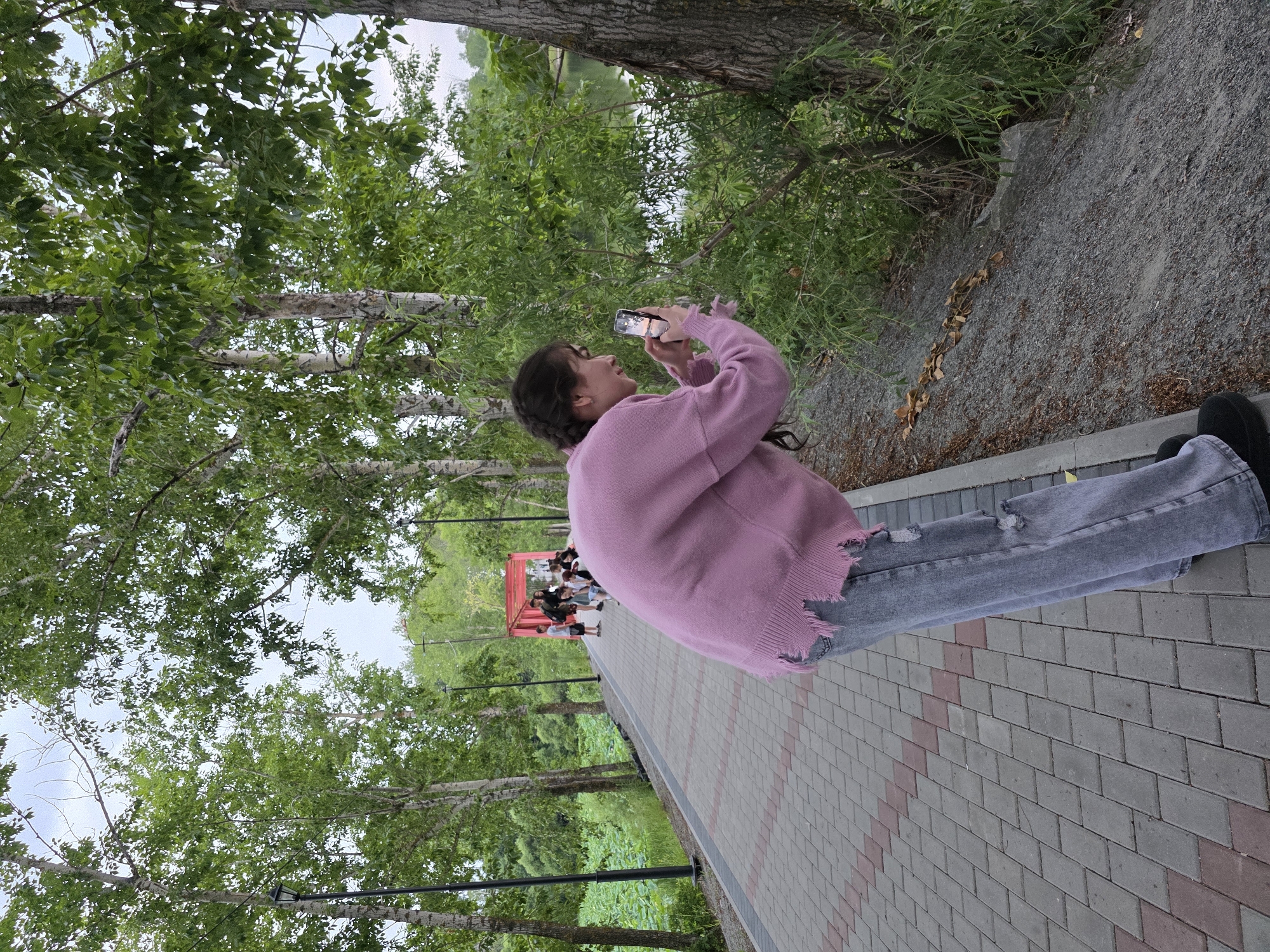 A young woman with braided dark hair, wearing a distressed pink knit sweater and ripped grey jeans, is captured from behind as she stands on a paved walkway beside a wooded area and a body of water. She is holding a smartphone in her hands, appearing to take a photograph or video of something to her right. The walkway is made of interlocking grey and red bricks. In the background, a group of people are visible near a bright red pedestrian bridge. The environment suggests a park or recreational area, likely located in Progress, Russia, with numerous trees and lush green foliage. The lighting indicates it is daytime, possibly late afternoon given the angle of the sun casting light on the phone screen.