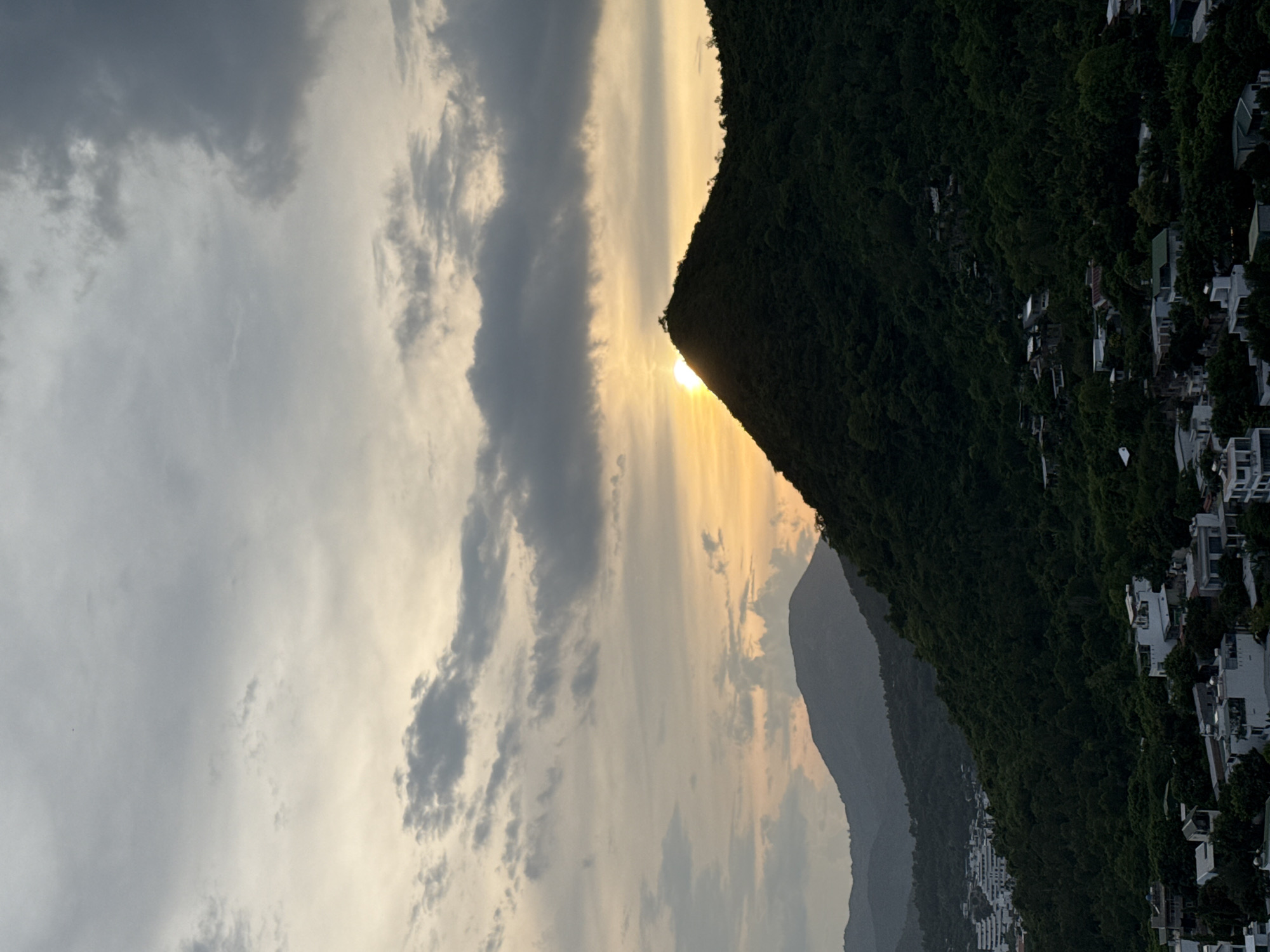 This image captures a scenic sunset over a lush, mountainous landscape dotted with buildings. The sun, a bright orb, is partially obscured by the peak of a dark green, tree-covered mountain, casting a warm, golden glow that extends across the sky. Wispy clouds, tinged with yellow and orange hues, are scattered across the sky, indicating a dramatic sunset.
The foreground is dominated by the silhouette of the densely forested mountain, its slope receding into the distance. Nestled on the mountainside and spreading along the lower slopes are numerous white buildings, their modern architecture contrasting with the natural environment. These structures suggest a residential area or perhaps a resort, built into the hillside. In the distance, further mountain ranges are visible, fading into a hazy, muted gray, likely due to atmospheric conditions and the fading light.
The time of day is clearly sunset, evidenced by the low position of the sun and the warm, atmospheric lighting. The weather appears to be clear enough for a sunset, though some clouds are present. The location context provided places this scene in the Bac Natural Heritage Area, Trang, Vietnam, a detail that hints at a potentially natural and protected environment where human development is integrated into the landscape. The overall mood evoked is one of tranquility and natural beauty, enhanced by the dramatic play of light and shadow. There is no visible text in the image.
