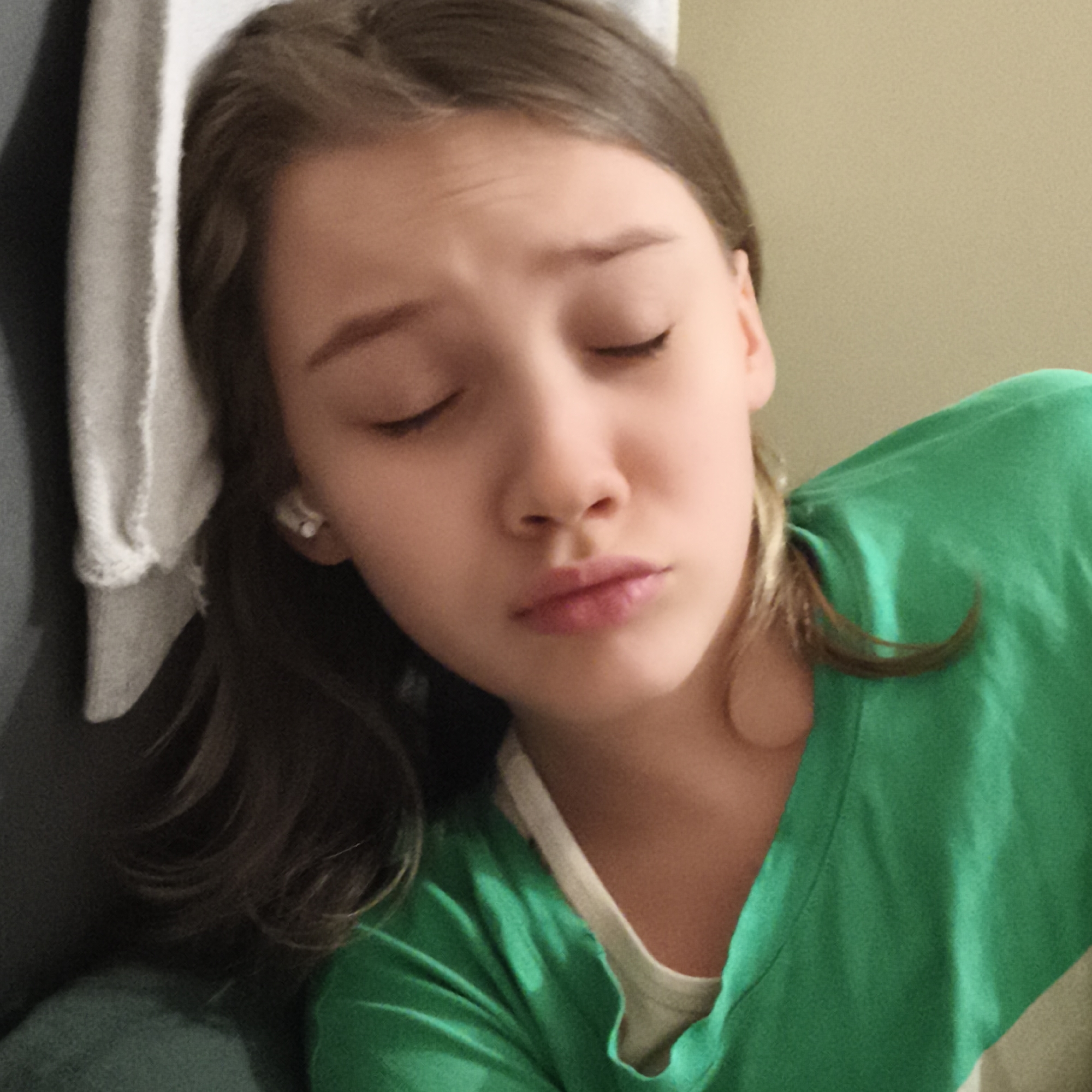 A young person with closed eyes is wearing a green shirt and lies against a gray surface next to a white towel. Their hair is brown, and they appear to be resting. The background is a plain wall of unknown color. The location is Unknown, Unknown.