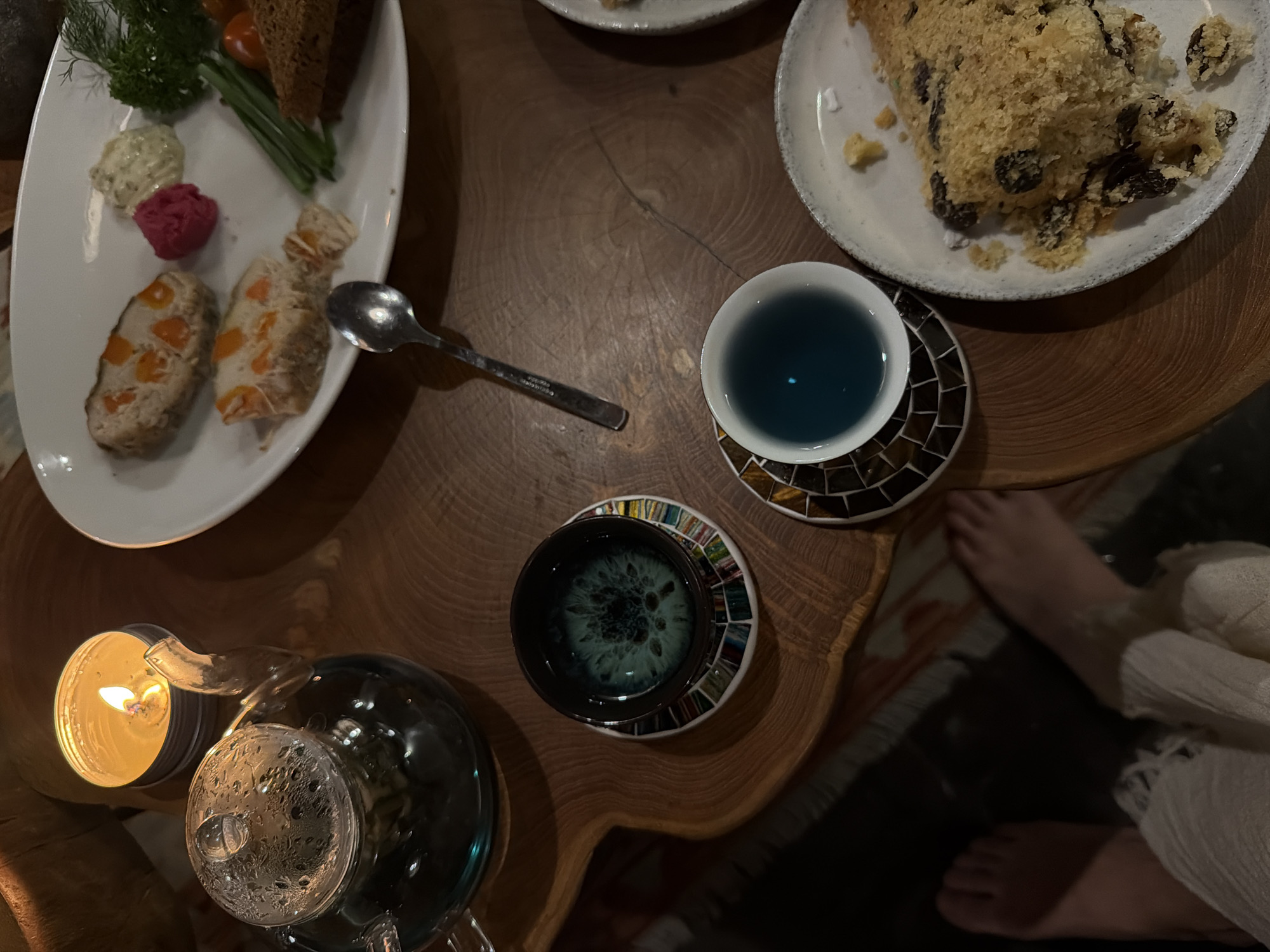 The image depicts a wooden table set with various items. A white plate holds appetizers including bread, spreads, and vegetables. Another white plate contains a slice of cake with raisins. Two cups, one containing blue liquid and the other a dark green liquid, sit on mosaic coasters. A silver spoon lies between the plates and cups. A glass teapot and a lit candle are visible at the lower left. In the lower right, a person's bare feet and a portion of their white clothing are visible. The scene likely takes place in Bongkasa, Indonesia, although this cannot be verified from the image alone.