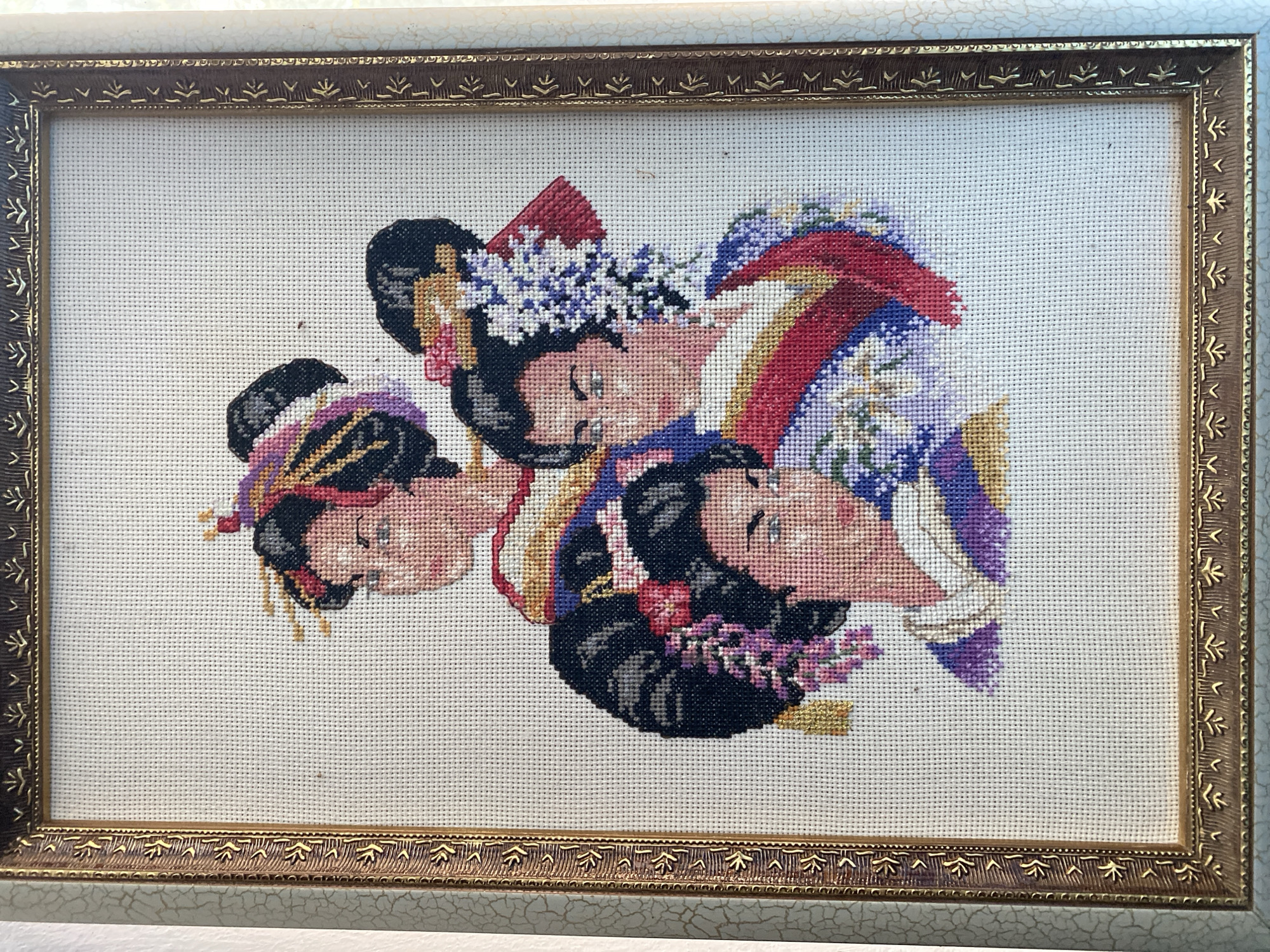 The image displays a framed cross-stitch artwork featuring the portraits of three women in traditional Japanese attire, depicted from the chest up. The women have dark hair styled elaborately with ornaments and flowers. Their faces are rendered with delicate features and subtle expressions. They wear colorful kimonos with intricate patterns and bold red sashes. The background is a plain white fabric, characteristic of cross-stitch. The frame is ornate, with a textured, aged-gold finish and decorative patterns along its edges. The artwork is hung on a wall with a light, textured wallpaper. The scene is indoors, suggesting a domestic or display setting. The location is unspecified.