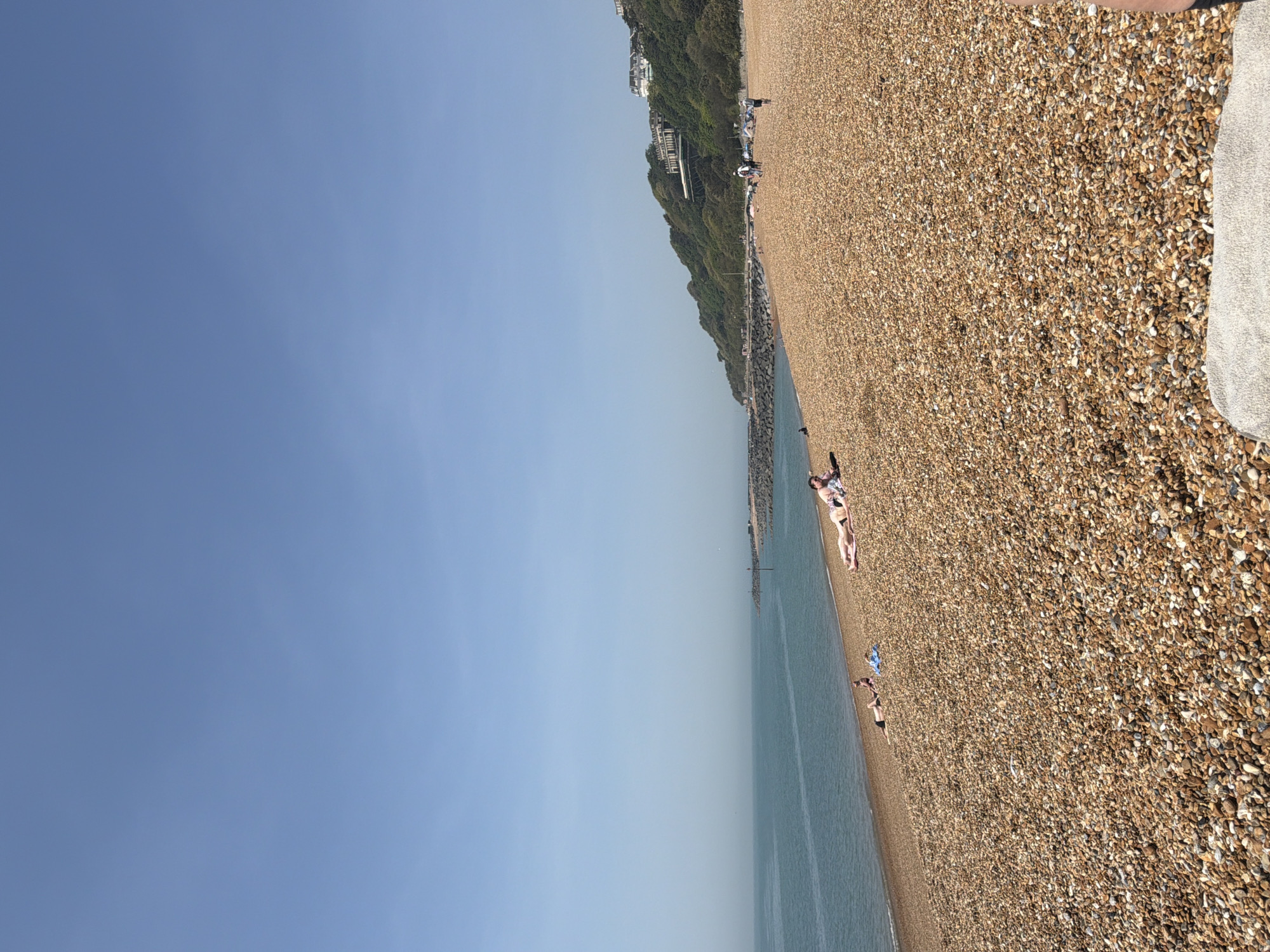 The image shows a sunny day at a pebble beach in the Folkestone and Hythe District, United Kingdom. The sky is predominantly blue. A body of water, likely the sea, runs along the length of the beach. The shoreline is covered in small pebbles and stones. Several people are on the beach, some sunbathing and others possibly walking. A green, overgrown hillside rises adjacent to the beach, and some buildings are visible near the top of the hill. A towel or fabric object rests on the pebbles close to the foreground.