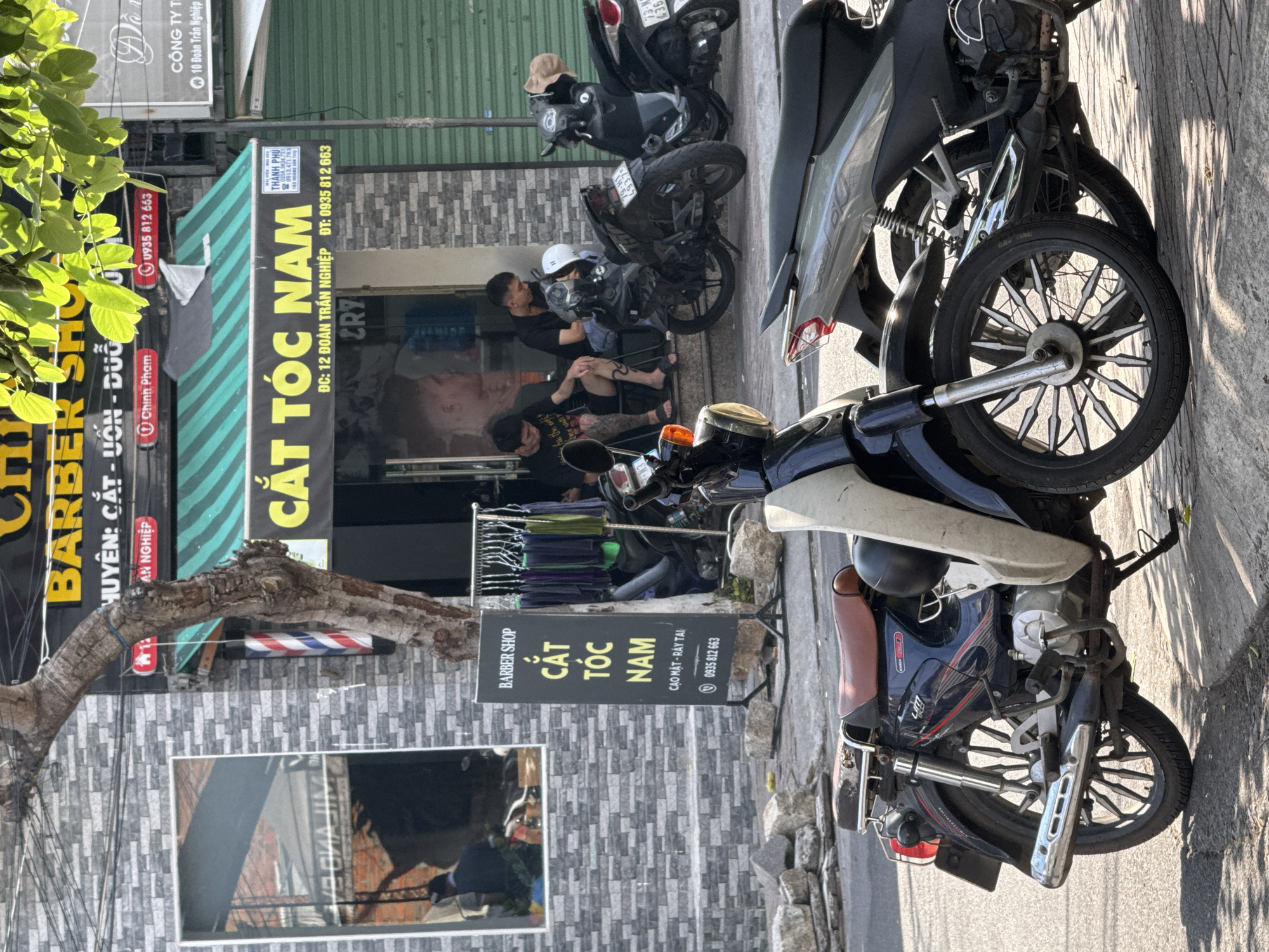 The image depicts the exterior of a barber shop in Bac Nha Trang Ward, Vietnam, with two men sitting on the steps outside and several motorcycles parked on the sidewalk. The barber shop, identified by signage reading "BARBER SHOP CẮT TÓC NAM" (Barber Shop Men's Haircut) and "HUYỆN: CẮT - UỐN - DUỖI" (District: Cut - Perm - Straighten), features a green striped awning. One man, wearing a black t-shirt and shorts, has tattoos on his legs. The other man, in a black t-shirt, is looking down. A white helmet sits on a black motorcycle. A tall, dark tree with a barber pole on its trunk stands to the left of the entrance. To the right of the entrance, green metal shutters are visible. A black sign on an easel in front of the shop displays "BARBER SHOP CẮT TÓC NAM CAO MẶT - RÂY TAI 0935 812 663". The sidewalk is paved with gray tiles, and several grey stones are scattered on the ground. In the foreground, a dark blue motorcycle with a brown seat is parked, with a lighter colored motorcycle to its right. A reflection in the mirrored building to the left shows an interior with red brick walls and people.