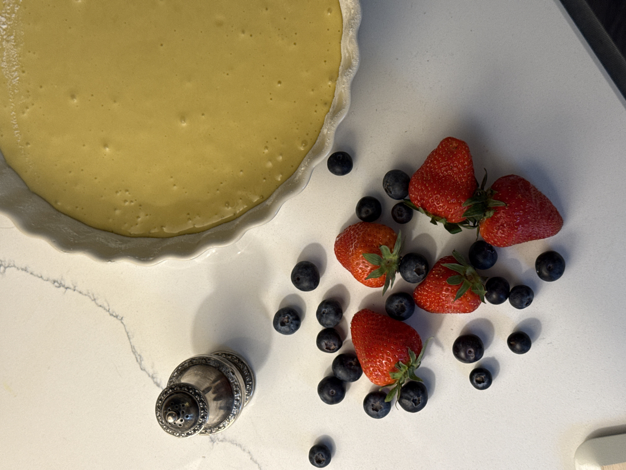 A top-down shot shows a partially-filled pie crust, strawberries, blueberries, and a small silver object sitting on a white countertop. The pie crust is in the upper left of the frame. It appears to be unbaked and contains a yellow liquid filling. To the right of the pie are five strawberries and a scattered arrangement of blueberries. In the bottom left is a short, round silver object with a patterned surface. The countertop has faint grey veining, and a dark, out-of-focus edge is visible in the upper right corner. There are no people visible. This image does not contain information about the location in the city of Moscow, Russia.