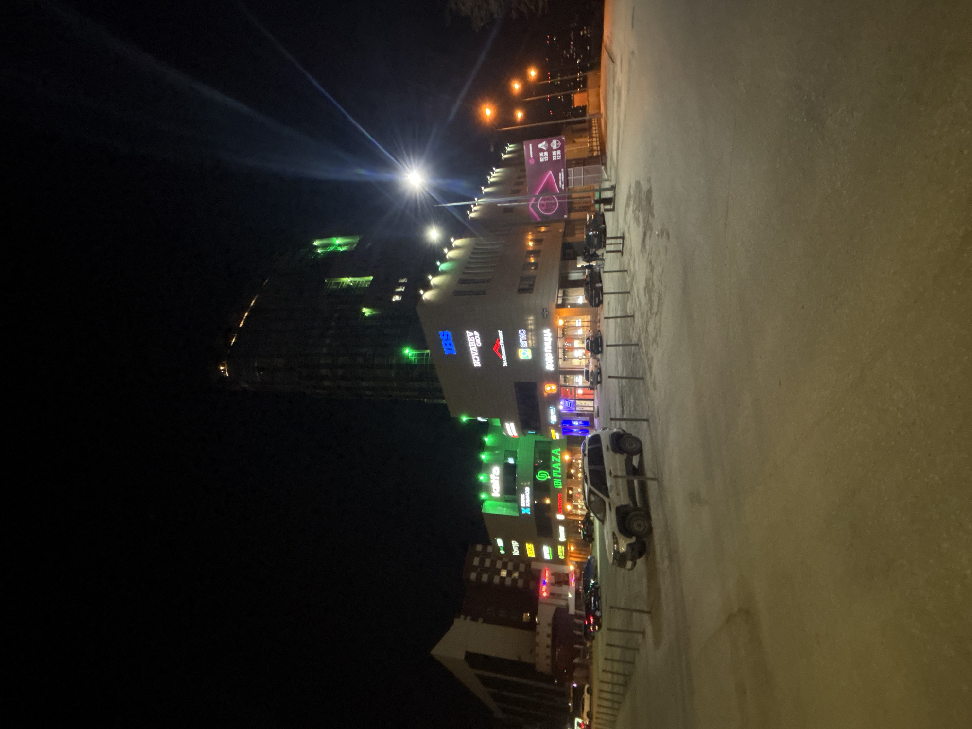 The image depicts a night scene in Perm, Russia. The primary subject is a multi-story building complex adorned with illuminated signage, including "SBS Megamall," "Novazakaz Group," "Uralchemtrans," and "додо Пицца." A taller structure is visible in the background, featuring rows of lit windows. Various smaller businesses and restaurants are indicated by neon signs. The foreground contains a paved area, partially obscured by low-light conditions. Several parked cars are visible along the right side of the frame, near a row of bicycle racks. A bright overhead light source illuminates the scene, creating lens flare and highlights on the buildings and pavement.