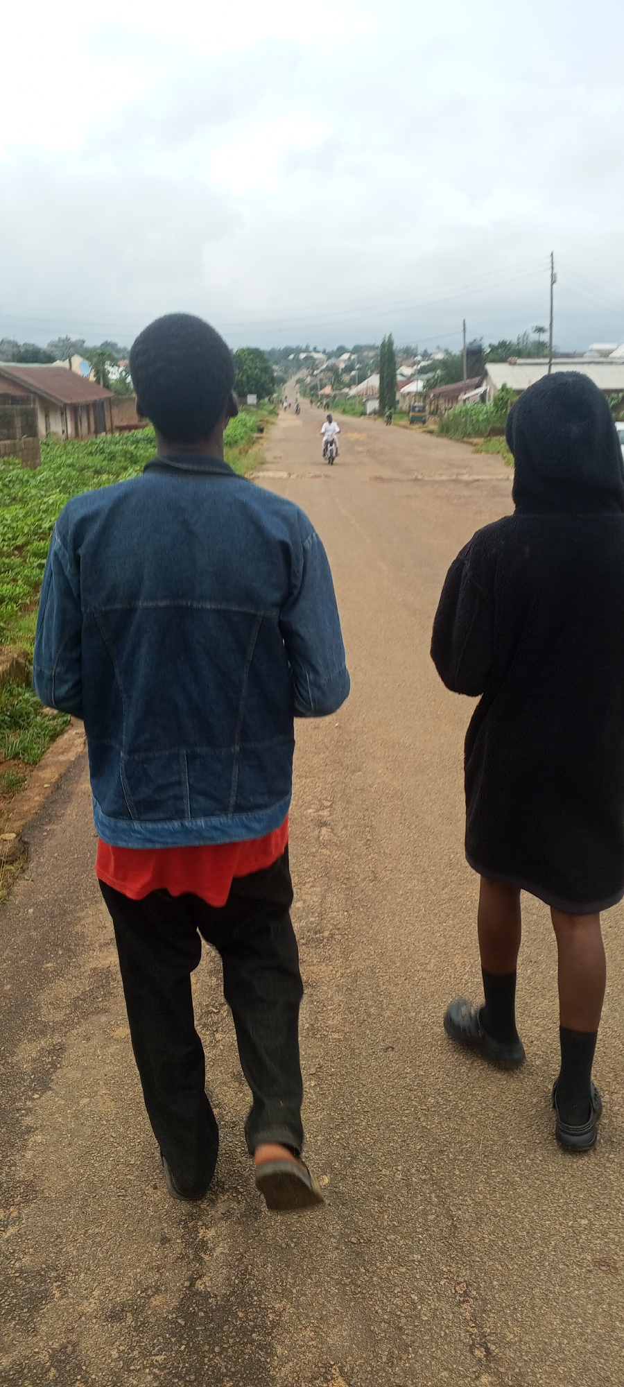 The image shows a street scene in Bahit, Nigeria, on a cloudy day. Two individuals are walking away from the camera on a paved road. The person on the left wears a denim jacket over a red shirt and dark pants. The person on the right wears a black hooded garment and dark socks with clogs. In the background, houses and vegetation line the sides of the road, and a person on a motorcycle is visible further down the road. The overall environment suggests a rural or semi-urban setting.