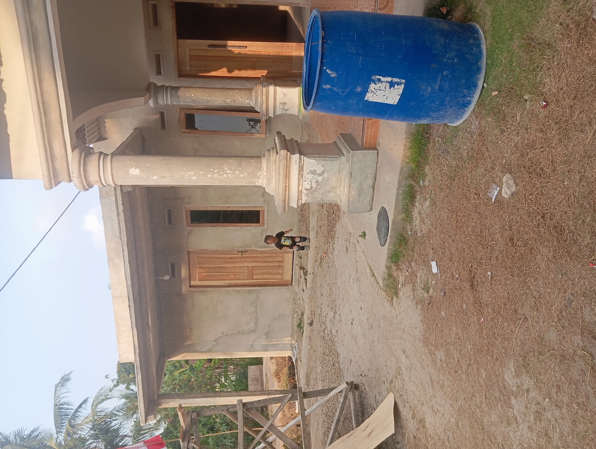 A young child stands in front of a house under construction in Dente Teladas, Indonesia. The house has unfinished concrete walls and wooden doors and windows. A large blue plastic barrel sits on the right side of the image. A wooden scaffold is visible on the left. The ground is covered in dirt, gravel, and dry grass. The sky is clear and blue.