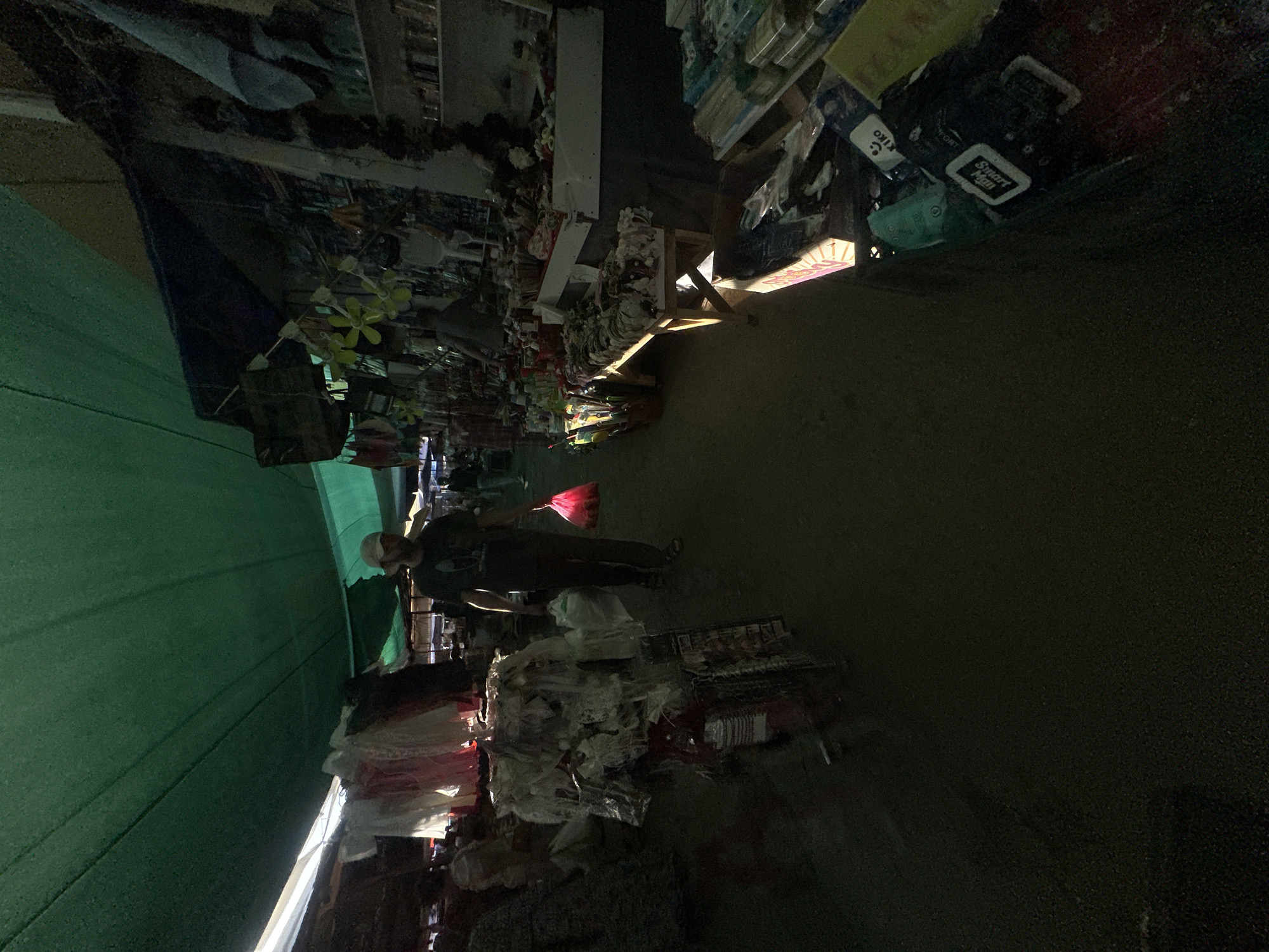 A man walks through a market under a green awning, holding a pink plastic bag. The market appears to be outdoors, with stalls selling various goods on either side of a dirt path. Some stalls are covered with tarps. The man is wearing a dark t-shirt, dark pants, and a light-colored baseball cap. He is walking away from the camera. The aisle is filled with merchandise, including clothing, bags, and what appear to be small toys or decorative items. The lighting is dim, with some areas illuminated by sunlight filtering through the awning and some artificial light. The environment suggests a vibrant, busy marketplace, possibly in Tashkent, Uzbekistan, given the context.