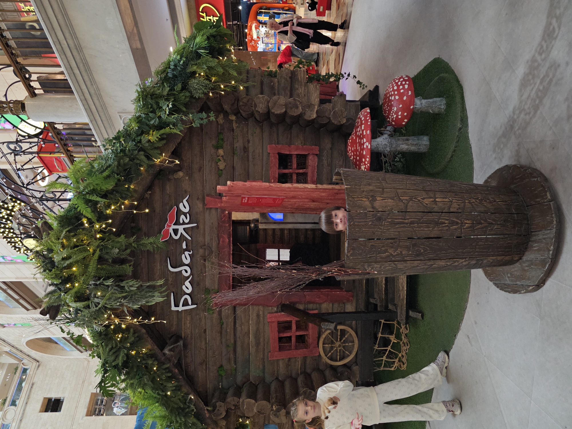 The image depicts a whimsical, fairy-tale-themed installation in a public indoor space, likely a shopping mall, in Moscow, Russia. At the center is a rustic, log cabin structure adorned with lush greenery and twinkling fairy lights, suggesting a festive or themed exhibit. The name "Баба-Яга" (Baba Yaga) is written in stylized white lettering above the cabin's entrance.
In the foreground, a young girl with blonde hair in a ponytail stands to the left, wearing a white sweater and pants with a sparkly pattern, and sneakers. She is looking towards the camera and has her hands extended as if holding or throwing something small. To the right of the cabin, a young boy with brown hair peeks out from behind a large, wooden, barrel-like structure.
Surrounding the cabin are decorative elements like large, red and white fly agaric mushrooms, and a cluster of dried branches. A wooden cartwheel is visible on the side of the cabin. In the background, other shoppers can be seen, including a woman in a pink scarf and black outfit, and a person in a red shirt. The overall environment suggests a commercial or entertainment venue, possibly during a holiday season or special event. The flooring appears to be light-colored tiles.