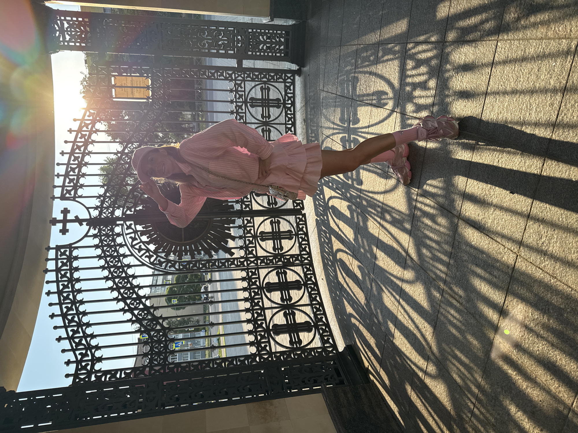 A woman in a pink outfit poses in front of a large, ornate black metal gate in Kirishi, Russia. The sun is setting, casting long shadows on the paved ground. The gate features intricate scrollwork and circular designs with crosses. In the background, a street with traffic lights and buildings is visible through the gate. The woman is wearing a pink collared shirt, a ruffled skirt, pink socks, and pink sneakers, along with a pink baseball cap. She has one hand on her hip and the other raised to her forehead, as if shielding her eyes from the sun. A small, quilted handbag hangs from her shoulder.