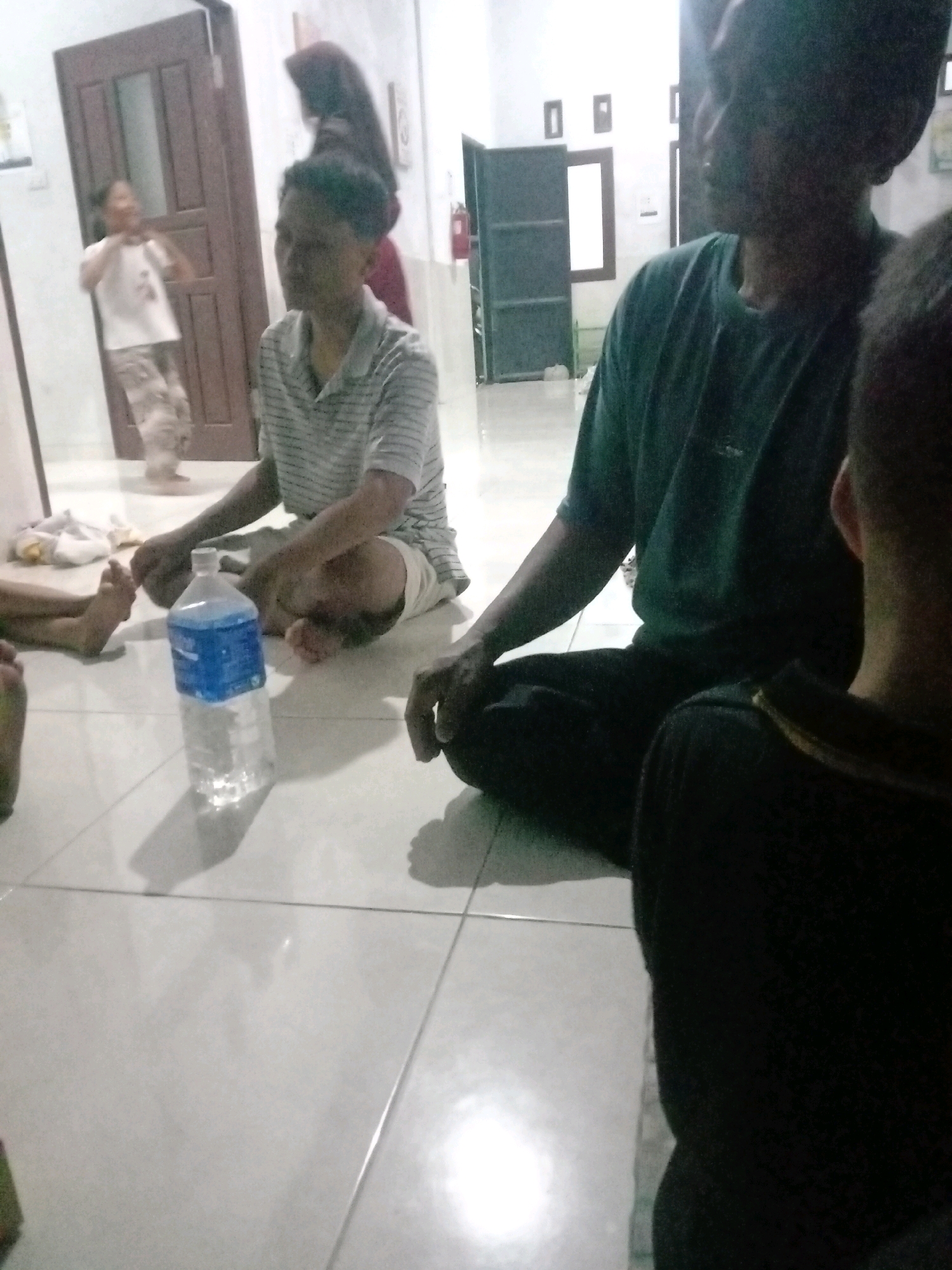 In the city of Rejosari Mataram, Indonesia, an indoor scene captures several individuals seated on a tiled floor. A person wearing a striped shirt and shorts is prominently positioned in the mid-ground, facing left. To their right, another individual is partially visible, wearing a dark green t-shirt and dark pants, facing towards the left foreground. In the background, a person wearing a white t-shirt and patterned pants is visible near a doorway. Another person, partially obscured and wearing a dark head covering, is situated behind the individual in the striped shirt. A plastic water bottle with a blue label is placed on the floor between the two individuals in the mid-ground. The environment appears to be a room with a tiled floor and light-colored walls, with a dark wooden door visible on the left. Shelving units are also present in the background.