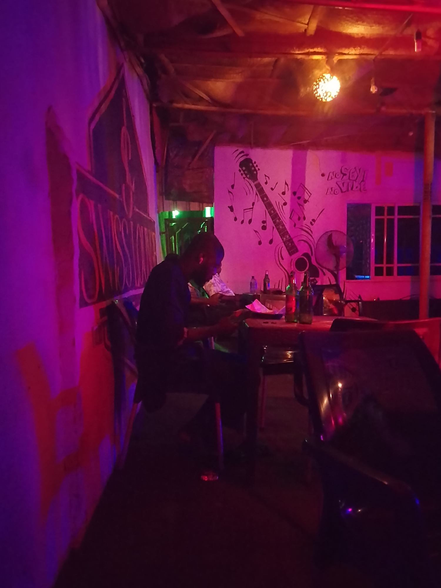 The image shows the interior of a dimly lit bar or restaurant with a man seated at a table. The location appears to be a casual establishment with unique wall art, including a painted guitar and musical notes. The atmosphere is relaxed, with a visible fan and soft lighting. The man seems engrossed in paperwork or reading, possibly working in the laid-back environment. Bottles of drinks are visible on the table, indicating that it is a bar setting. The location is in the city of Unknown, Unknown.