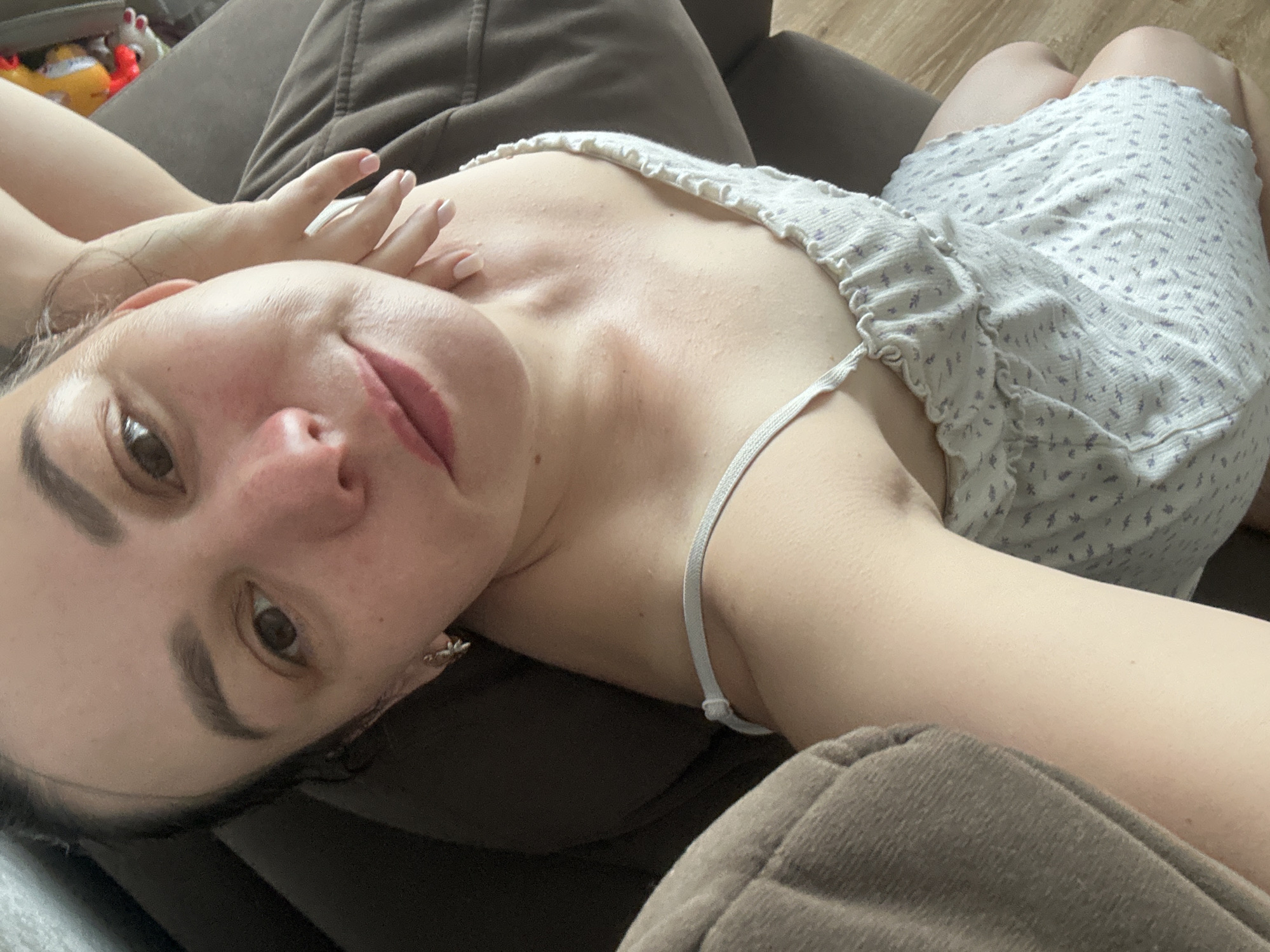 A selfie, taken indoors, shows a person with light skin lying on a dark-colored sofa. The person is wearing a light-colored patterned dress or top with thin straps. Their left arm is raised, with their hand near their face. The face is angled towards the camera, revealing brown eyes, eyebrows, and lips. The background includes part of the sofa and a hint of wooden flooring. The location is unknown and is not implied by the image. There is no data on people, subjects, objects, actions, or interactions in Chaikovsky, Russia.