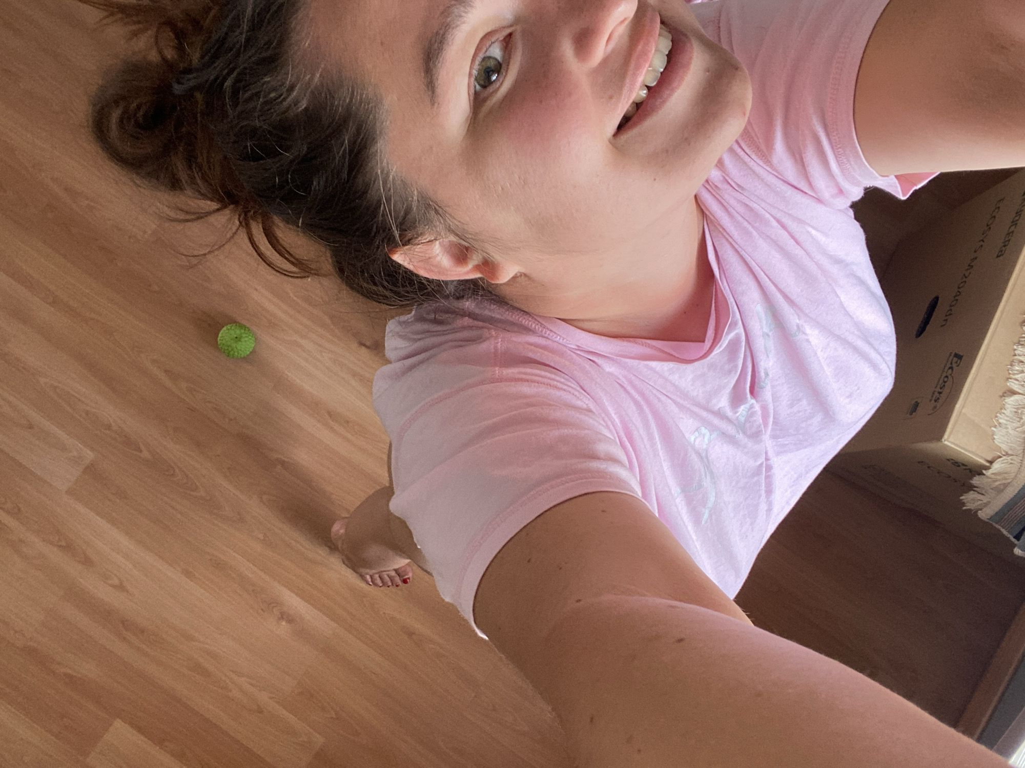 The image shows a close-up, high-angle selfie of a woman in a pink t-shirt. She has her hair pulled up in a messy bun and is smiling at the camera. Her left foot, with red nail polish, is visible in the lower left corner. A small, bright green ball is on the wooden floor to her right. The background consists of a wooden floor and what appears to be the edge of a cardboard box. The lighting suggests an indoor environment, possibly a home. There is no indication of the specific location within the city of Perm, Russia, nor any information to infer it.
