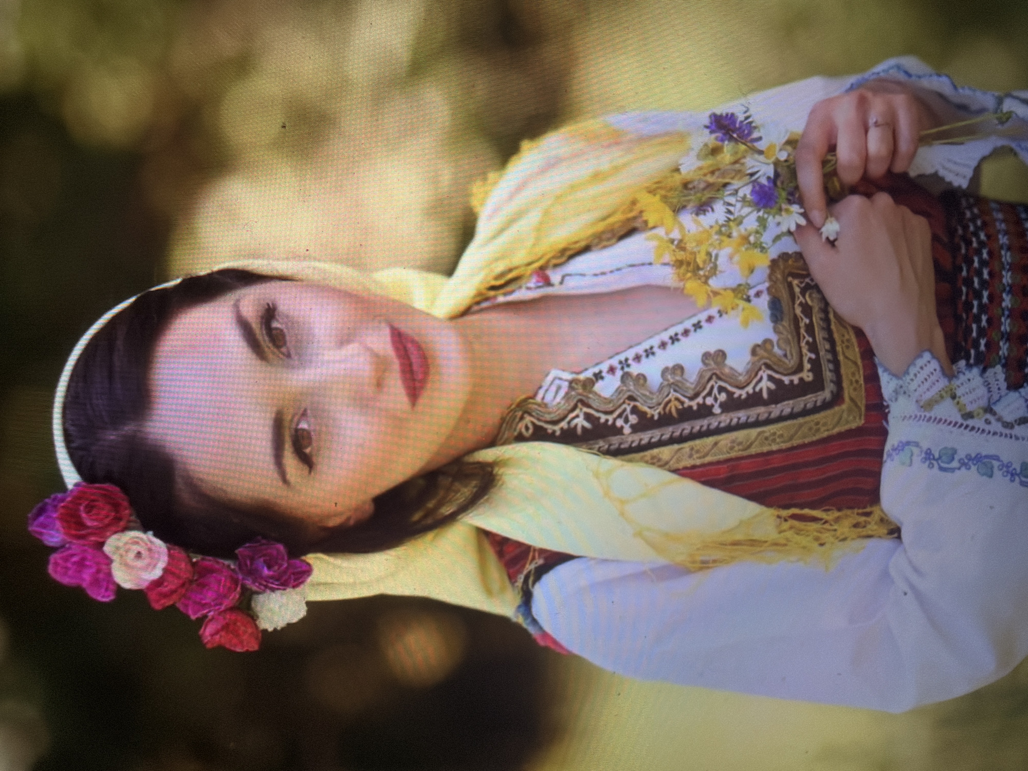 A young adult female is depicted in a full-frame portrait, positioned outdoors in the city of Shumen, Bulgaria. She faces directly towards the viewer with a slight smile. Her dark hair is adorned with a white headband and a cluster of artificial flowers in shades of red, pink, magenta, and white on the left side of her head. A light yellow scarf with fringed edges is draped over her head and shoulders. She wears a white long-sleeved blouse with intricate blue, white, and purple embroidery on the cuffs and along the front placket. Over the blouse, she has a traditional vest or bodice featuring dark brown fabric with gold and cream embroidery around the edges, and a central panel of vertical red and white stripes accented with patterned bands. Her hands are clasped at chest level, holding a small bouquet of wildflowers, primarily yellow, with additional white and purple florets. A silver-colored ring with a clear stone is present on her left ring finger. The background is a blurred, out-of-focus natural environment composed of green and yellowish-brown tones, indicative of foliage under soft daylight.
