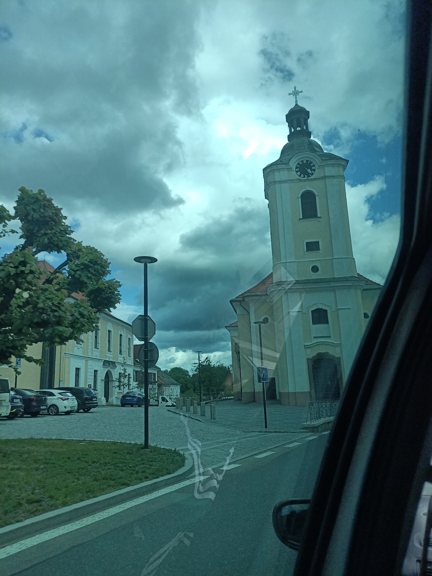The image captures a street scene in Dalovy, Czechia, featuring a prominent church under a partially cloudy sky. The photograph is taken from inside a vehicle, evidenced by the dark car door frame visible on the far right and reflections on the lower portion of the windshield.
At the center-right, a light-colored church dominates the view, featuring a tall square tower topped with a dark, ornate belfry and a cross. A clock with Roman numerals (XII, I, II, etc.) is visible on the tower, indicating approximately 12:50 PM (short hand just past XII, long hand on X). The church facade is a pale yellow or cream color with white architectural details around windows and corners. Below the tower, the church building has a reddish-brown tiled roof and multiple arched and rectangular windows, some with dark shutters. A dark, ornate door is visible at the ground level of the church.
To the left of the church, a paved area, likely a plaza or square, features cobblestones. To the left of this area, a patch of green grass curves alongside a curb that borders an asphalt road. Several vehicles are parked on the cobblestone area to the left, including a white hatchback, a dark blue sedan, and a black SUV. Further down the street, another blue car is visible. A large deciduous tree with dense green foliage stands prominently on the far left, partially obscuring a multi-story building with a light-colored facade and a red-tiled roof.
A tall, black lamppost with two circular signs attached stands on the edge of the cobblestone area near the center-left. The sky is largely overcast with significant dark grey clouds, but patches of bright blue sky are visible, particularly behind the church tower, suggesting dynamic weather conditions. No people are discernible in the scene.