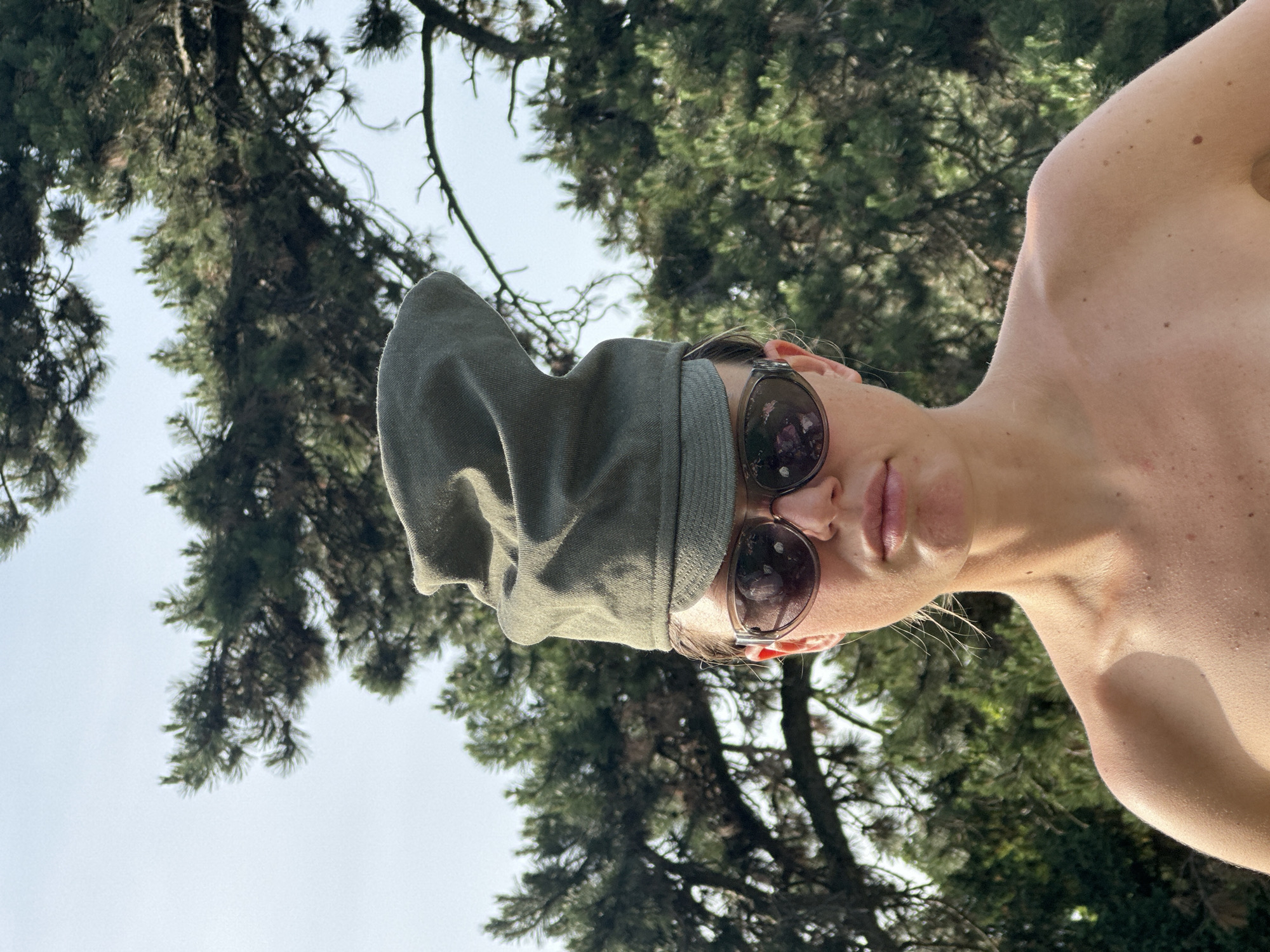 A selfie, taken at a low angle, shows a person with pale skin wearing sunglasses and a green hat. The person's face is angled towards the camera. The individual is partially nude, with their shoulder visible. The background shows a bright sky and green pine trees, suggesting an outdoor setting. It is not possible to confirm that this photo was taken in Paris, France.