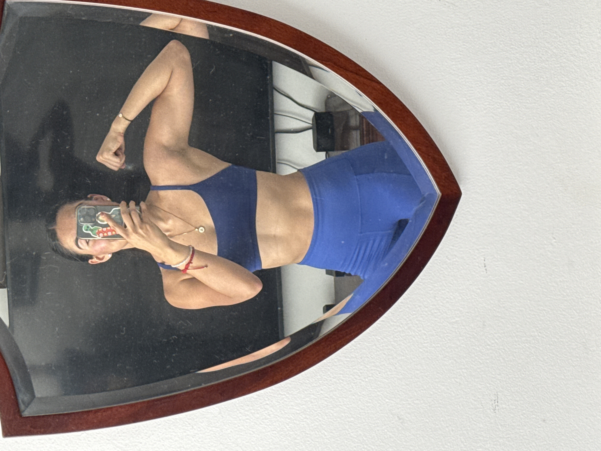 A person in blue athletic wear is reflected in a mirror. The mirror has a dark wood frame and is positioned against a white wall. The person is holding a smartphone, presumably taking a photograph or video in the mirror. The background, visible in the mirror, includes electrical wiring and possibly a portion of a room. There's no discernible information to indicate the location is Puebla, Mexico.