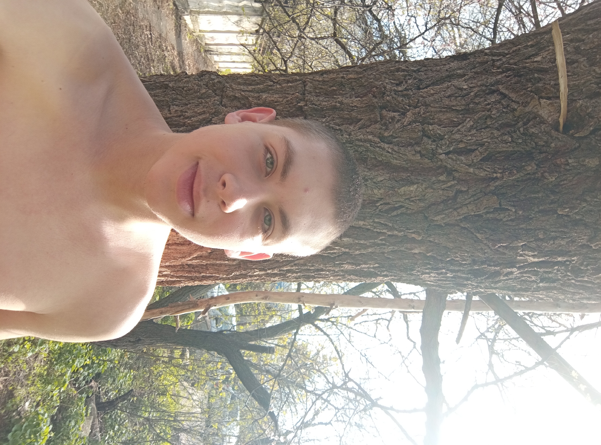 This image captures a young man outdoors in what appears to be a natural or semi-natural setting, possibly in совхоза "Воронежский", Russia. The photo is oriented ninety degrees clockwise. When viewed upright, a young man, shirtless with short light-brown hair and striking light-green eyes, is prominently featured on the left side of the frame. He looks directly at the camera with a calm, subtle smile, his face illuminated by bright sunlight which causes some glare on his skin.
To his right, a large tree trunk with rough, textured bark dominates the middle and right side of the composition. The man appears to be standing very close to or leaning against this tree. In the background, a bright, clear sky is visible through a dense network of bare tree branches, suggesting a time of year when deciduous trees are without leaves, possibly early spring or late autumn. Some faint green foliage can be seen in the lower background, along with a glimpse of an indistinct fence or structure in the upper left background. The strong sunlight suggests it is daytime, casting bright highlights across the scene. There is no visible text.