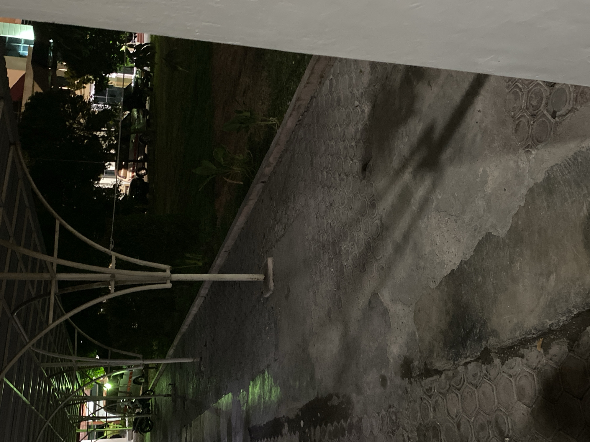 The image depicts a covered walkway at night in an urban environment. The walkway is paved with interlocking hexagonal blocks and has a white metal structure supporting a roof overhead. The ground is wet and reflects the green light from an unseen source. To the left, a motorcycle is parked under the walkway. Beyond the walkway, there is a grassy area with trees and several vehicles, including a van and what appears to be a car. In the background, a building with illuminated windows is visible. Shadows are cast on the walkway, indicating artificial lighting. The overall scene suggests an outdoor parking area or a public space at night.