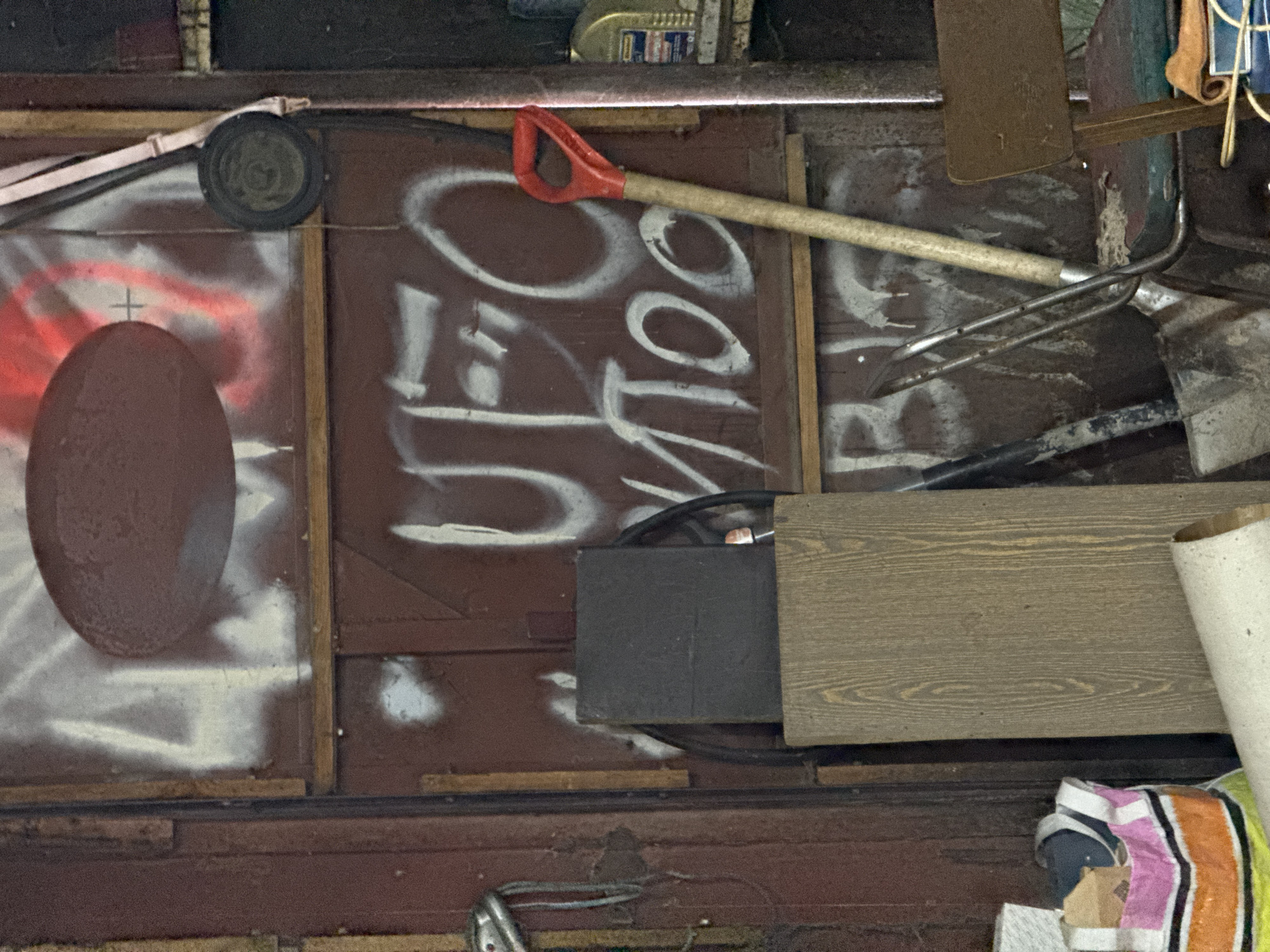 The image shows a cluttered storage space, likely a shed or garage, in Naro-Fominsk, Russia. Various tools and objects are piled up, including a shovel with a red handle, a garden fork, a rectangular dark gray box, and a piece of wood with a prominent grain pattern. The background is a dark brown wooden wall covered in white spray paint graffiti, some of which appears to be lettering. A large oval shape, possibly a painted representation of an egg, is also visible on the left side of the wall, along with red spray paint strokes. A small wheel with a strap is mounted above the oval. The overall impression is one of disorganization and disuse. There is no indication of a specific time of day or weather conditions. The focus is on the accumulated items and the graffiti on the wall.