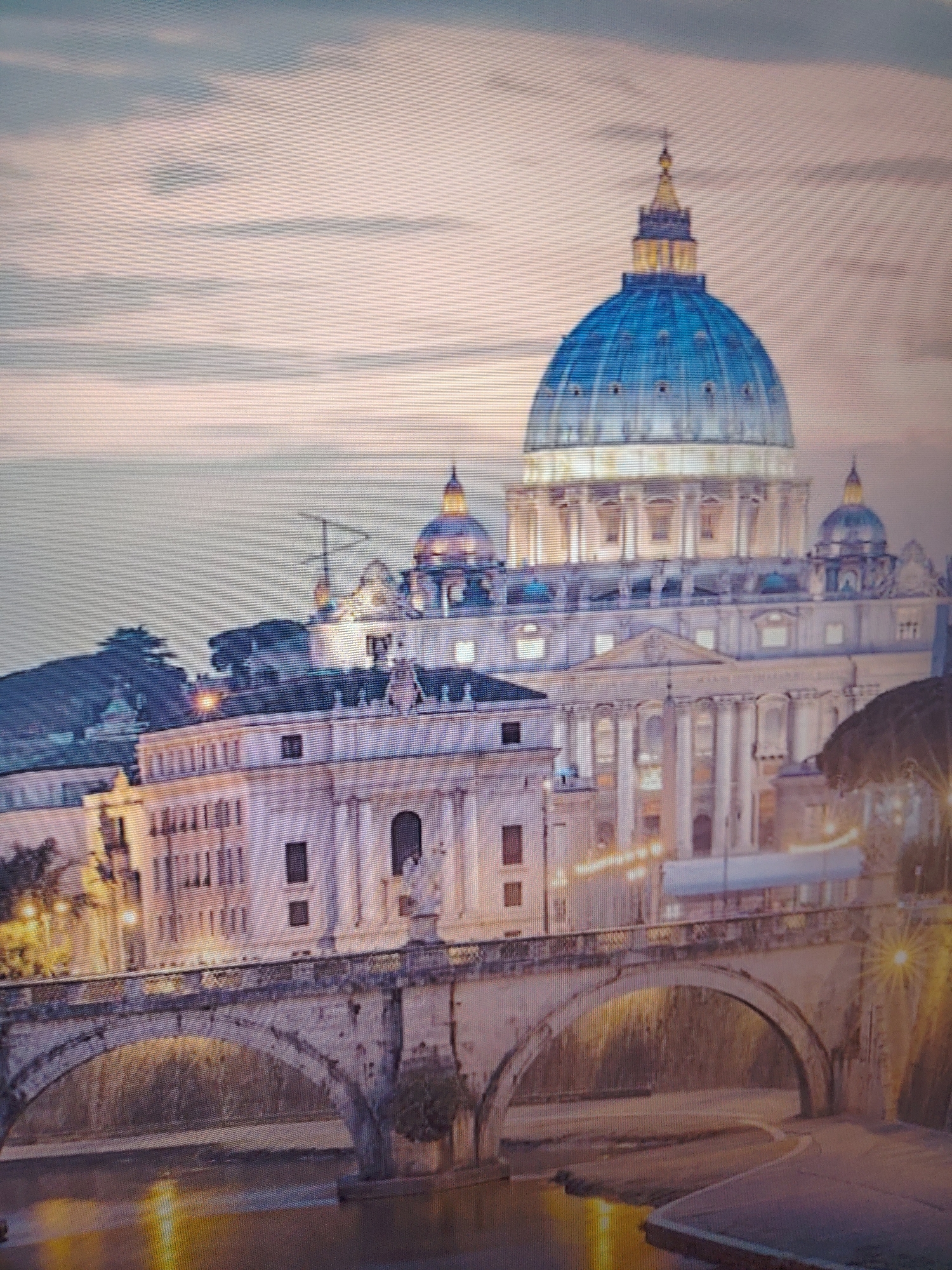 The image depicts a nighttime view of St. Peter's Basilica in Vatican City, Rome, Italy. The basilica's dome is illuminated and stands prominently against a cloudy sky. In the foreground, a stone bridge with arches spans a river, with light reflecting off the water's surface. The buildings around the basilica are also lit up, contributing to the overall illumination of the scene. No people are visible.
