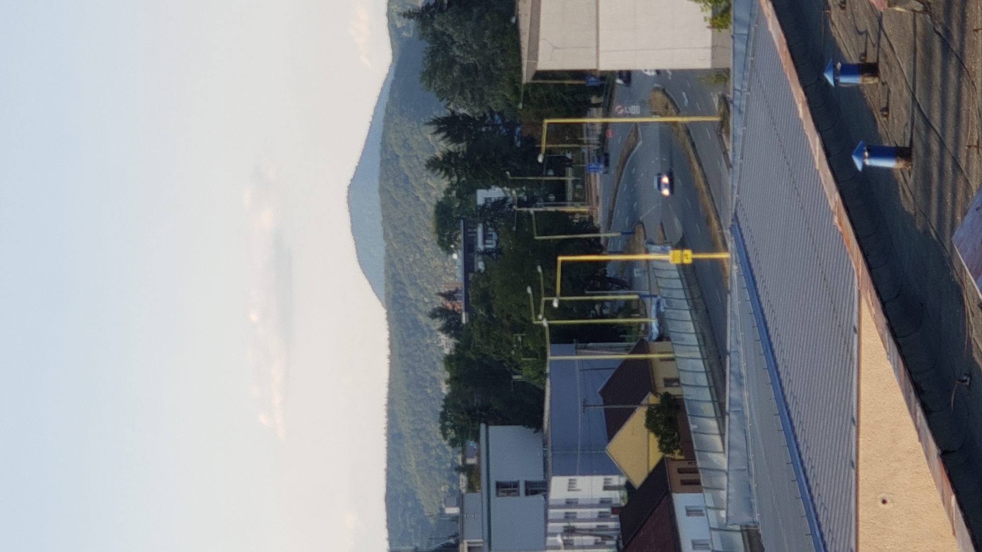 The image depicts a view of a city street in Prešov, Slovakia, on a clear day. The foreground features the rooftops of several buildings, one with a corrugated metal roof and another with a darker, possibly tar-covered roof featuring two blue vent pipes with pointed caps.
In the mid-ground, a two-lane road curves through the scene, with a yellow dividing line and a concrete barrier along its left side. Several tall, yellow lampposts line the road. Cars are visible on the road, including one with its headlights on, and another in the distance. Adjacent to the road, there are several residential buildings, including a yellow house and a white building with multiple windows. Behind these buildings, a dense collection of trees forms a green backdrop.
Further in the background, rolling hills covered in dark green trees extend across the horizon, with a distinct, rounded mountain peak visible in the far distance. The sky is a pale blue with a few wispy clouds. The overall impression is a typical urban landscape with elements of nature on the periphery.