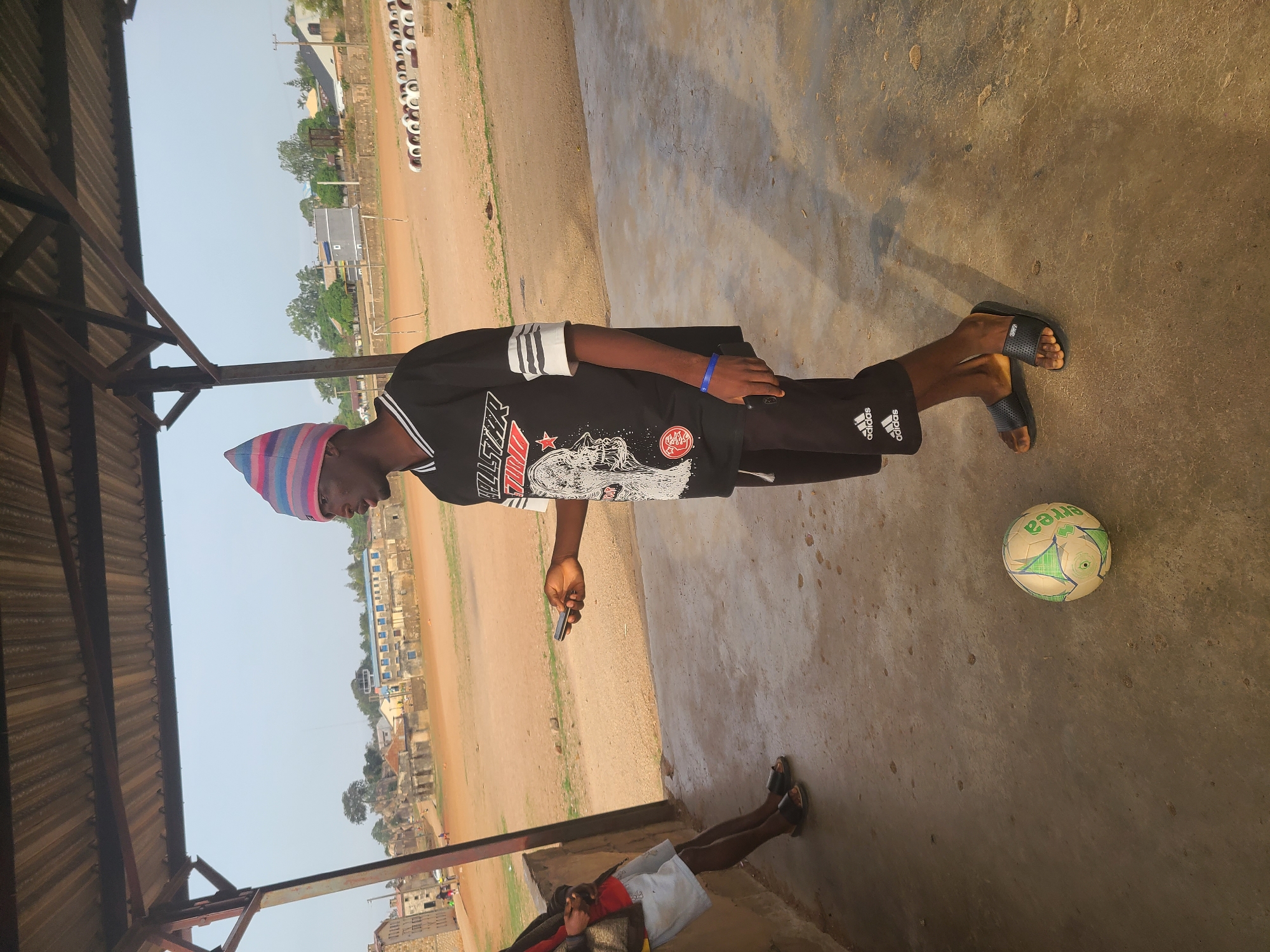 A person wearing a striped beanie, a black t-shirt with a graphic print, black shorts with Adidas logo, and sandals is standing on a concrete surface next to a soccer ball. He is holding a mobile phone. A second person is partially visible in the lower left corner. The setting appears to be a covered area with corrugated metal roofing. In the background, a field with a row of tires is visible, along with buildings and trees, indicative of an urban environment, likely Jos, Nigeria.