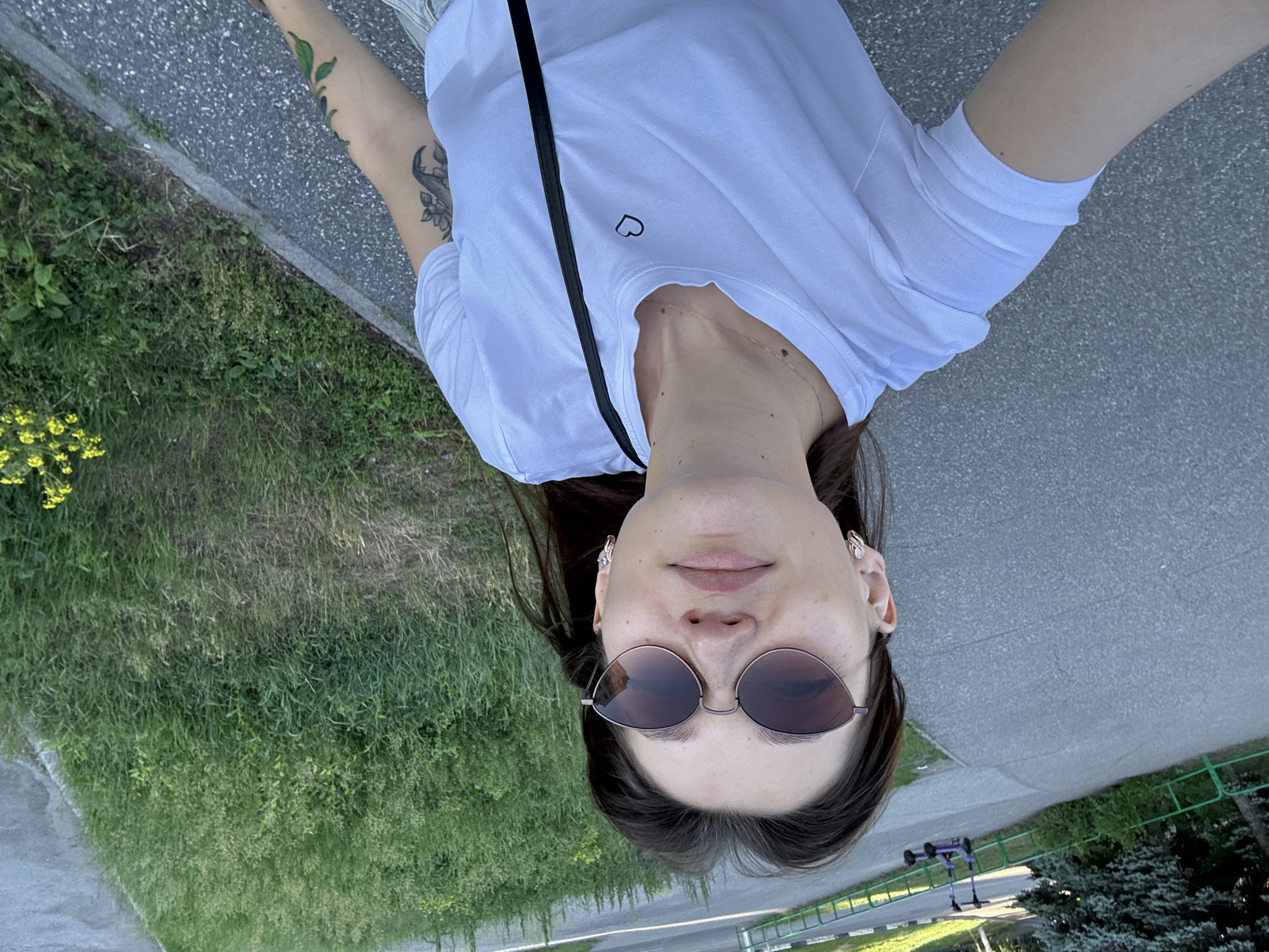 A young woman with long dark hair and sunglasses takes a selfie outdoors. She is wearing a white t-shirt with a small black heart graphic on the left chest and a black strap of a bag over her shoulder. She has earrings and a necklace. The background shows a paved pathway, a grassy area with yellow wildflowers, and trees. In the distance, there are two purple scooters parked. The scene appears to be a park or a recreational area in Novokuznetsk, Russia, possibly near a cycling path.