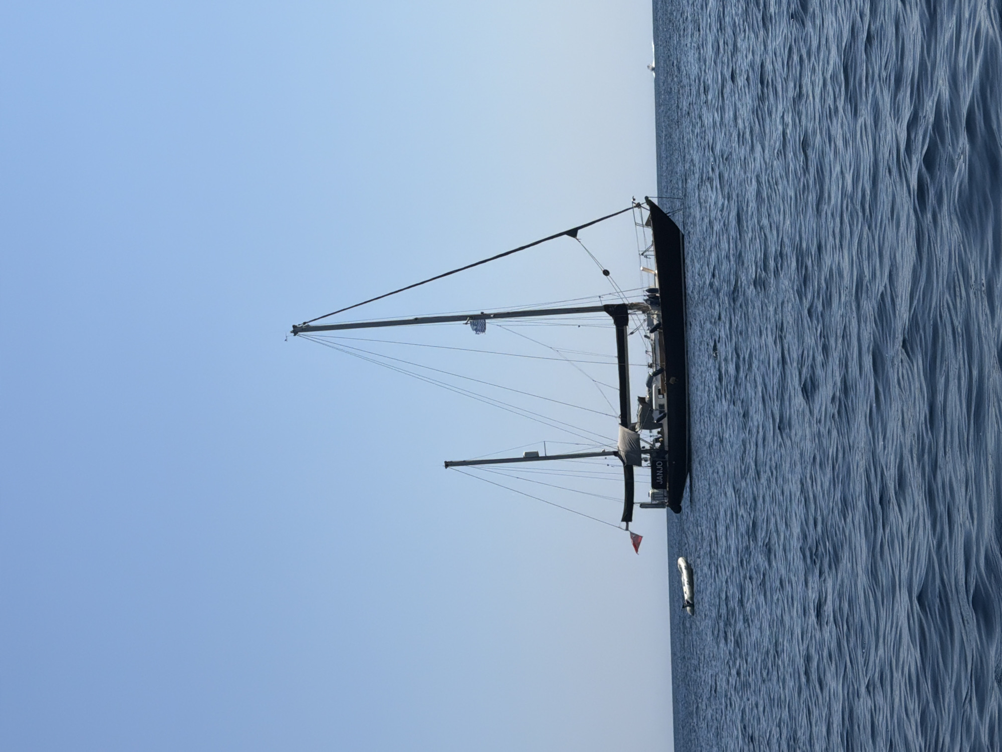An overview shot captures a large, dark-hulled sailboat with two masts anchored on a calm, blue sea under a clear, light blue sky. A small, white inflatable dinghy is visible in the water to the left of the sailboat. The name "JANJO" is visible on the stern of the sailboat. The distant horizon shows a faint line where the sea meets the sky, with a small white boat in the far distance. The scene appears to be set in the Municipal Unit of Kastos, Greece, a location known for its clear waters and popular boating destinations. The overall impression is one of tranquility and maritime activity.