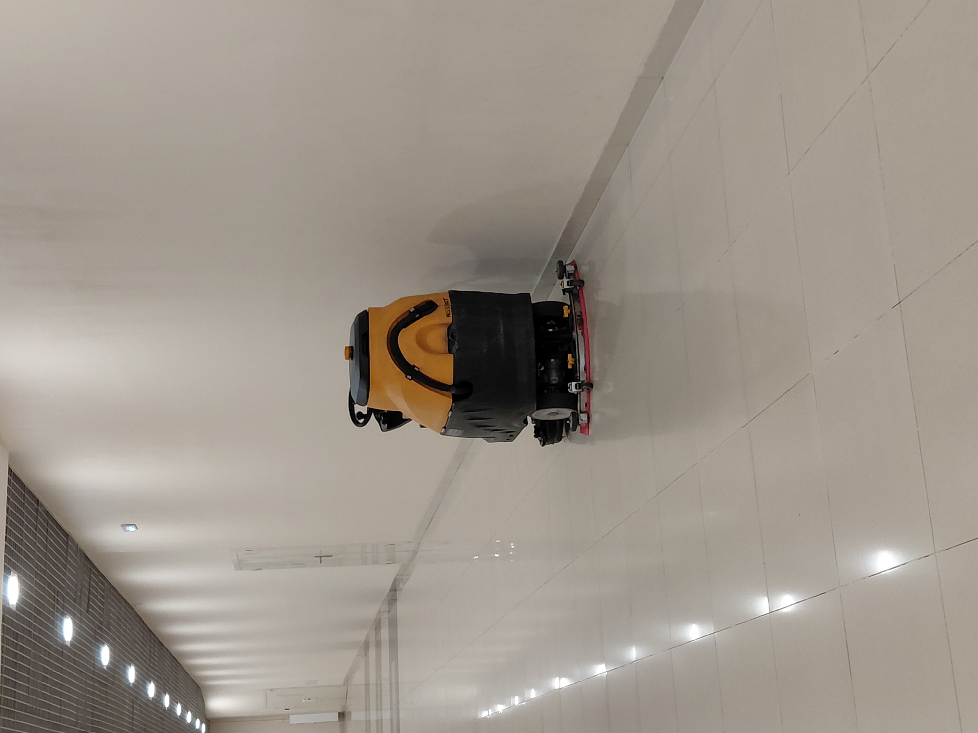 The media file shows a yellow and black floor cleaning machine in an indoor setting. The machine is positioned in the center of the frame and appears to be in operation, with its brushes visible. The floor is tiled in a light color, and there are some reflections on the surface. In the background, the ceiling is visible with recessed lighting. The scene appears to be a hallway or a large room with a tiled floor. There is no indication of the time of day or weather conditions, as it is an indoor environment. No people are visible in the frame. The machine has some text on it, but it is partially obscured and illegible. The overall impression is of a well-lit, clean, and modern indoor space.