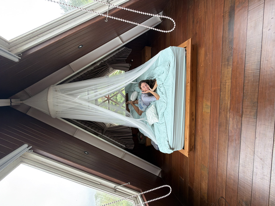 A woman lies in a bed draped with a white mosquito net in a room with wooden walls and floors. The bed is covered in light blue bedding and several pillows. The room features slanted ceilings with windows offering a view of greenery outside. White beaded cords hang near the windows. The woman is looking up towards the camera.