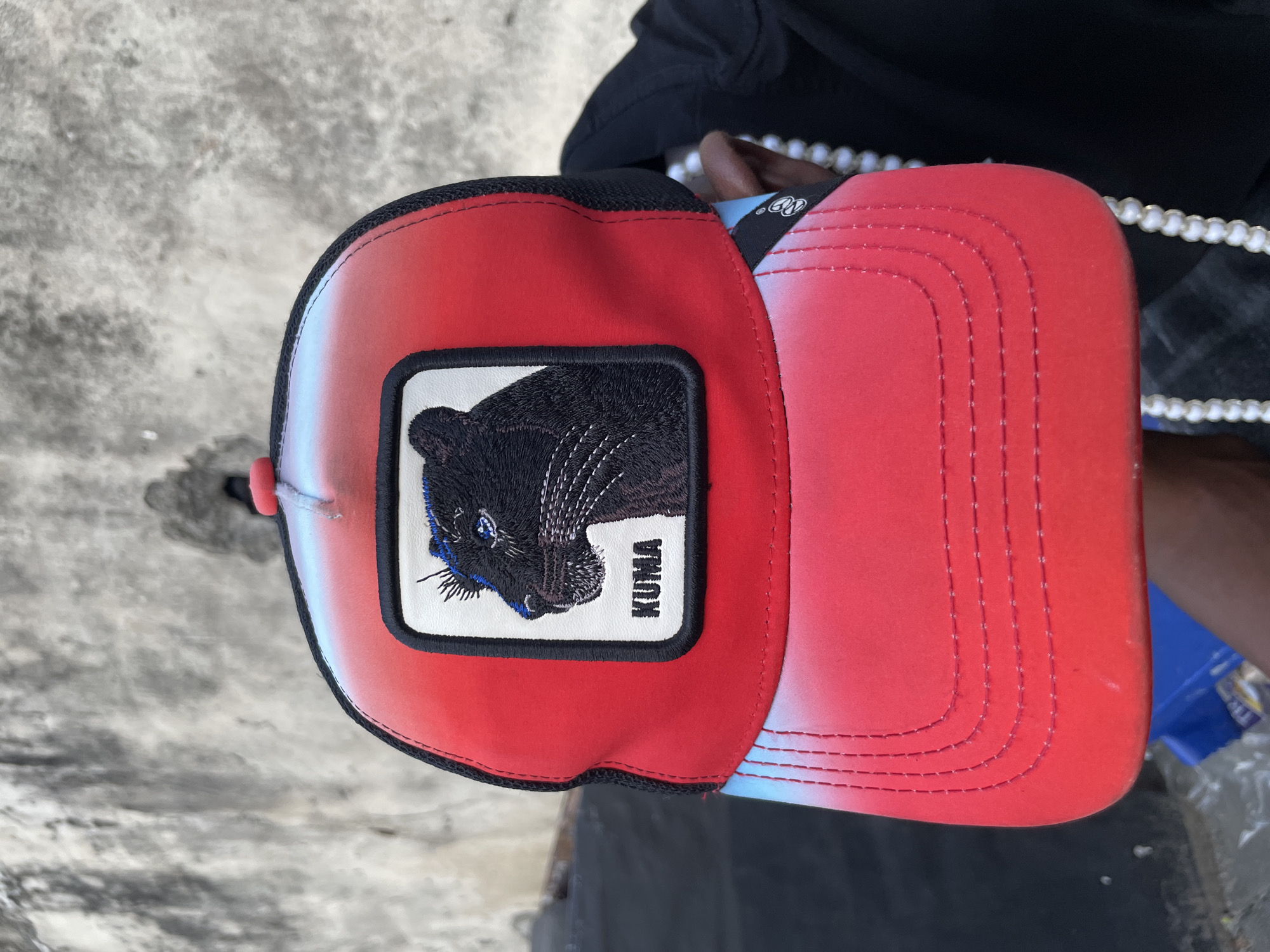 A close-up, eye-level shot shows a person wearing a red and gradient black trucker hat with a black panther patch. The patch is a square with a black panther head facing left, with the word "KUMA" in white text below it. The hat has a red front with a gradient of light blue and white towards the back, which is black mesh. The background is a textured concrete wall, likely in a street or outdoor setting in Ado, Nigeria. The person's dark skin is visible on their arm and ear, adorned with a pearl necklace. The focus is on the hat, with the background slightly blurred.