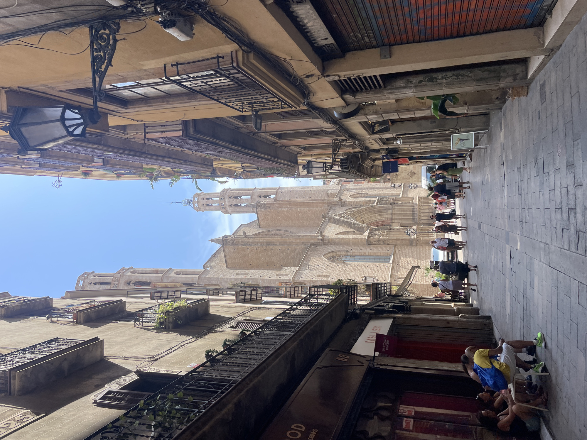 This image depicts a narrow street in Barcelona, Spain, with tall buildings lining both sides. A large, Gothic-style cathedral or church with prominent spires is visible at the end of the street. The buildings feature balconies with ornate wrought-iron railings and some have plants on them. Several people are walking down the street, and a few are sitting outside a shop on the left. The street is paved with stone. The buildings on the right have roll-down shutters, some of which are graffitied. The sky is clear and blue.