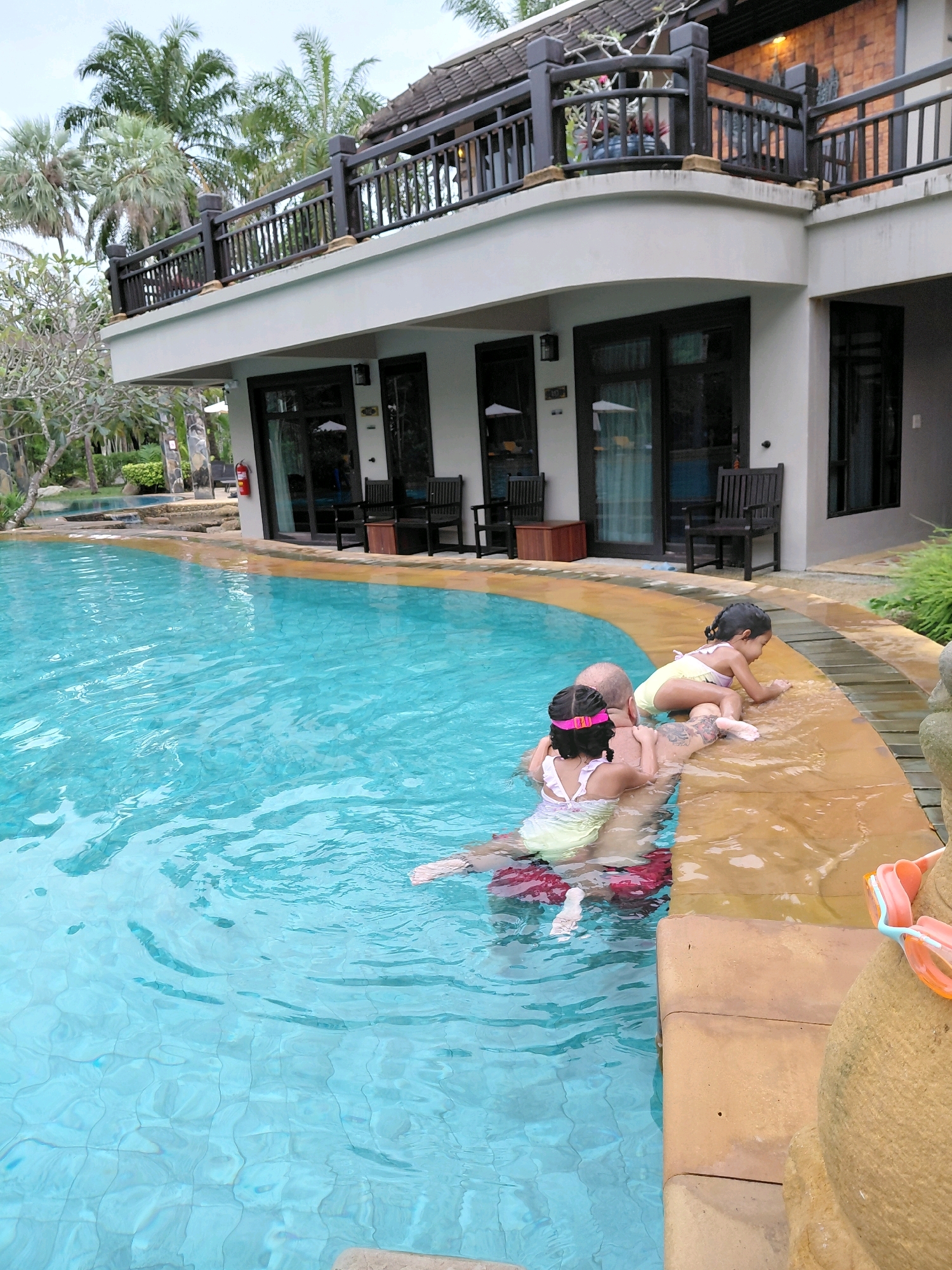 In a tropical resort setting in Ban Khuan, Thailand, a man is in a swimming pool with two young girls. The man is partially submerged in the clear blue water, and one girl is being held on his back while another is on the pool's edge. The older girl is wearing a yellow and white swimsuit, and the younger girl is wearing a pink and white swimsuit with a pink headband. They appear to be enjoying a playful moment in the water.
The resort features a multi-story building with balconies overlooking the pool, surrounded by lush palm trees and tropical vegetation. The building has a modern design with dark wooden railings and light-colored walls. The pool area is bordered by light brown tiling and a stone sculpture is visible on the right edge of the frame. Goggles are resting on the edge of the pool. The lighting suggests it is daytime, likely late morning or early afternoon, with bright, natural light. The overall mood is one of relaxation and family fun in a vacation environment. No visible text is present in the image.