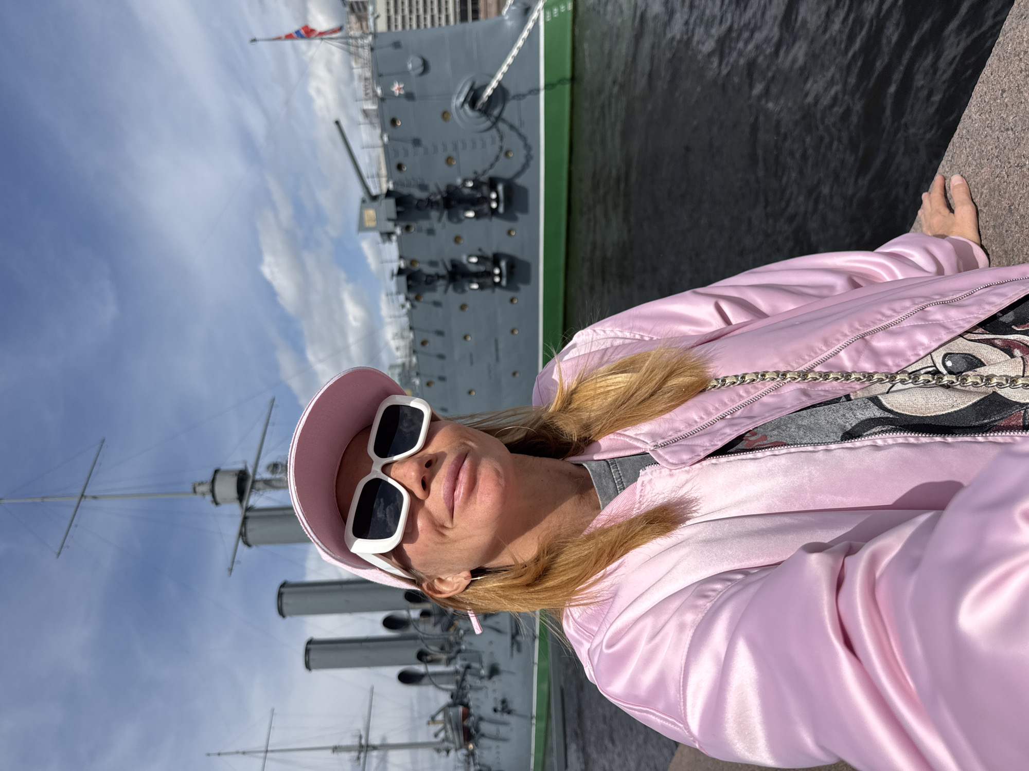 A woman takes a selfie outdoors in Saint Petersburg, Russia, with the historic Cruiser Aurora warship serving as her backdrop. The image is rotated 90 degrees clockwise.
In the foreground, the woman, with light brown hair, wears a vibrant pink satin bomber jacket, a matching pink baseball cap, and oversized white-framed sunglasses. A cartoon-style graphic is partially visible on her t-shirt underneath. She has a relaxed, subtle smile.
Behind her, the prominent grey Cruiser Aurora is docked on a body of water, likely a river or canal, alongside a stone embankment. The large warship features multiple gun turrets, one displaying a red star emblem, along with tall masts and smokestacks. A Russian naval flag (Andreevsky flag) flies from one of its masts. The water is dark and calm, with ripples.
The scene is set on a bright day under a partly cloudy sky, indicating favorable weather for sightseeing.
