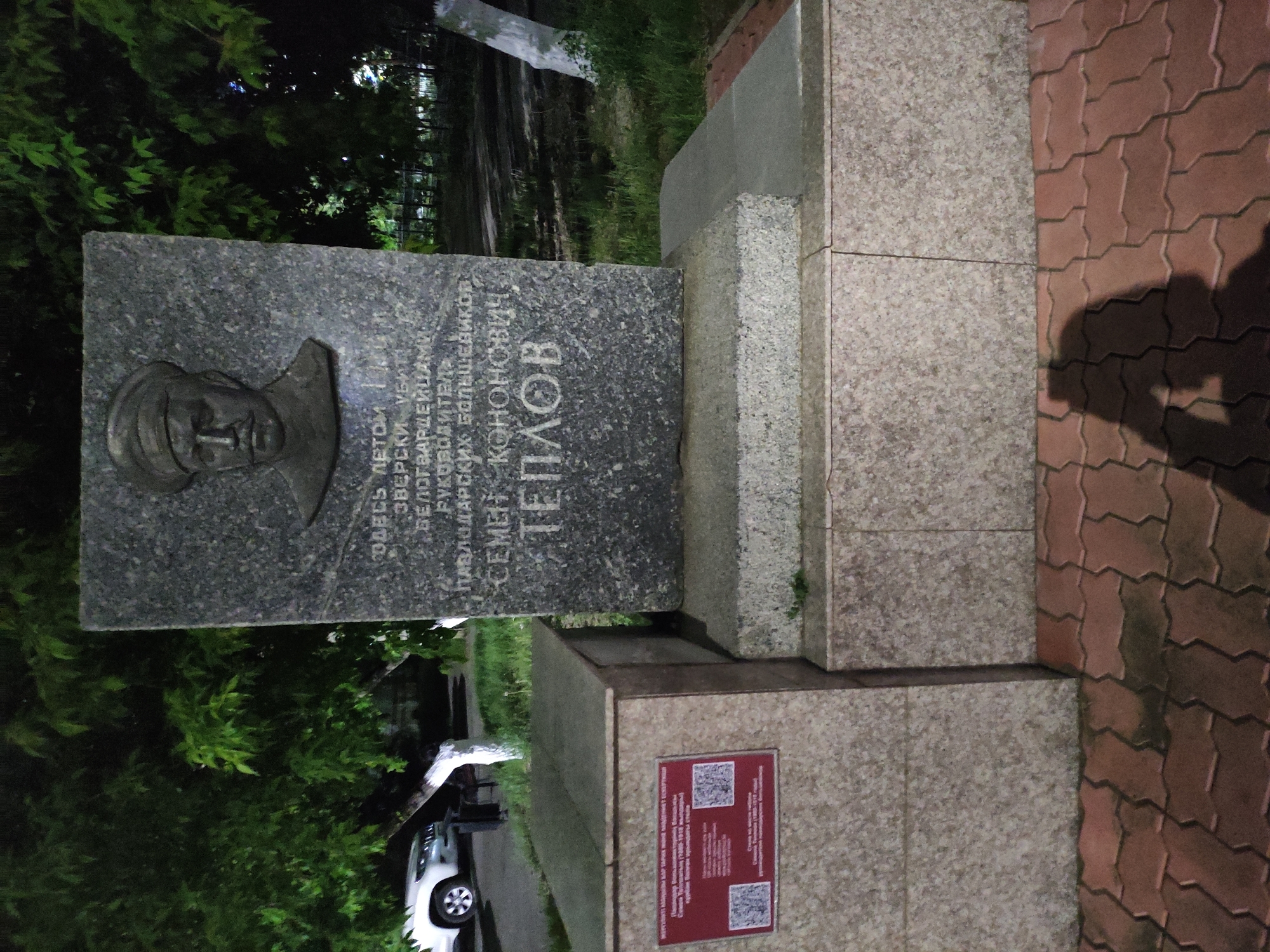 The image shows a monument in the city of Pavlodar, Kazakhstan, commemorating Semyon Kononovich Teplov, a leader of the Pavlodar Bolsheviks who was brutally murdered in the summer of 1918. The monument is made of gray stone and features a bas-relief portrait of Teplov. Inscription in Russian identifies him. The monument sits on a stepped base made of granite blocks and a QR coded sign is visible. The scene is lit artificially at night, and the shadow of a person taking the picture is visible on the bricked ground next to the monument. Trees are visible in the background, and a car can be seen on the left side of the image, implying its outdoor location is near a road.