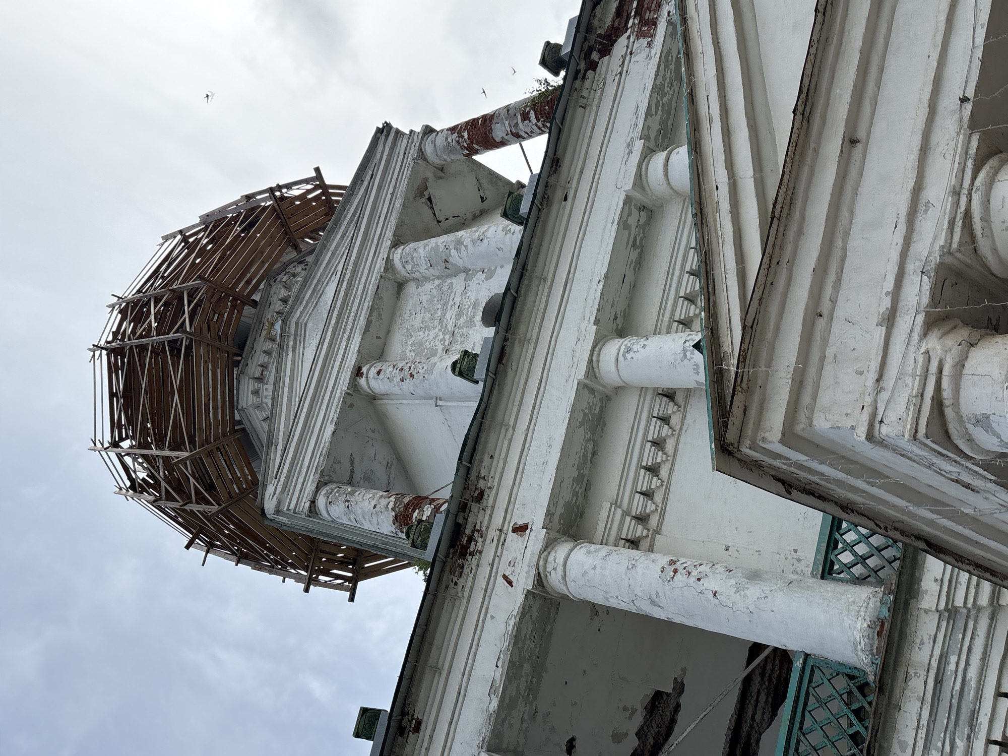 The image is a low-angle shot of the dome of a white, classical-style building that appears to be undergoing restoration. The dome is covered in scaffolding made of rough wooden planks. Below the dome, the building features white columns with weathered, red-brick sections and ornate white architectural details. The building shows signs of decay, with peeling paint and some visible damage. A turquoise-colored, lattice-like railing is visible on a lower level of the building. The sky above is overcast and pale. Two small birds are visible in flight in the upper right portion of the image. The building is identified as being located in the city of Kstinino, Russia.