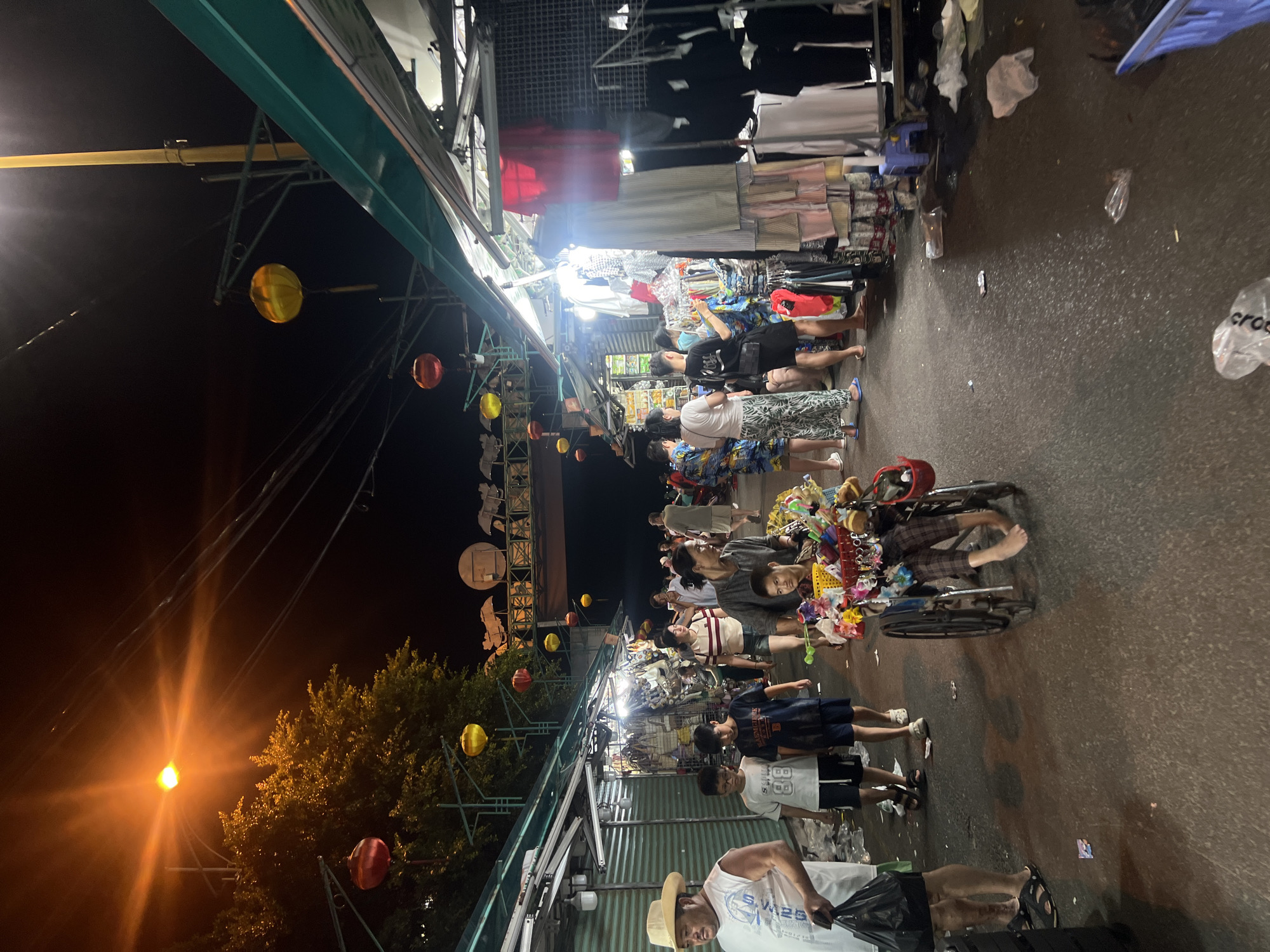 The image shows a crowded night market scene in Nha Trang Ward, Vietnam. People are walking along a narrow, paved street lined with stalls selling various items, including clothing and toys. Many are shopping. A person in a wheelchair selling trinkets is in the crowd. The stalls are covered with makeshift metal roofing supported by metal frameworks, decorated with hanging paper lanterns. The market is illuminated by bright overhead lights. Trees are visible in the distance, lit by a street lamp. Trash is scattered on the ground.