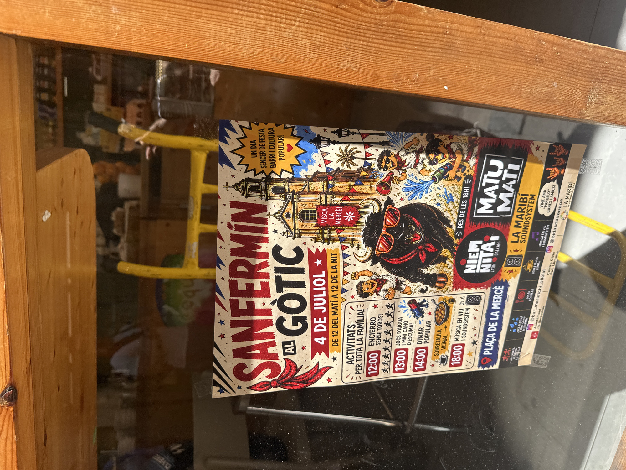 This is a photograph of a flyer posted on a glass surface, likely a shop window or door, in Barcelona, Spain. The flyer advertises an event called "SANFERMIN AL GÒTIC" taking place on July 4th from 12 PM to 12 AM. The event celebrates "a full day of festival, neighborhood, and popular culture."
The flyer features a colorful, illustrated design with a bull wearing sunglasses at the center, evoking a San Fermín theme. Other illustrations include people in traditional attire, a bull run with toy bulls, water games, a church-like building, and palm trees, suggesting a festive and family-friendly atmosphere.
The flyer lists various activities and their timings, such as "ENCICERRO SENSE TOROS!" (Bull run without bulls) at 12:00 PM, "JOCS D'AIGUA I MINI CANÓ D'ESCUMA!" (Water games and mini foam cannon) at 1:00 PM, and "DINAR POPULAR" (Popular meal) at 2:00 PM. It also mentions "MÚSICA EN VIU I SOUNDSYSTEM" (Live music and sound system) at 6:00 PM in "PLAÇA DE LA MERCÈ" (Mercè Square). Another section promotes "NIEM NITA! LATIN BALKAN!" and "LA MARIBÍ SOUNDSYSTEM".
The text is primarily in Catalan. The overall impression is of a lively community event designed to be enjoyed by all ages, blending traditional elements with modern entertainment. The lighting suggests it's daytime, with sunlight casting shadows. The background is blurred, showing the interior of a building with shelves and some yellow structures.