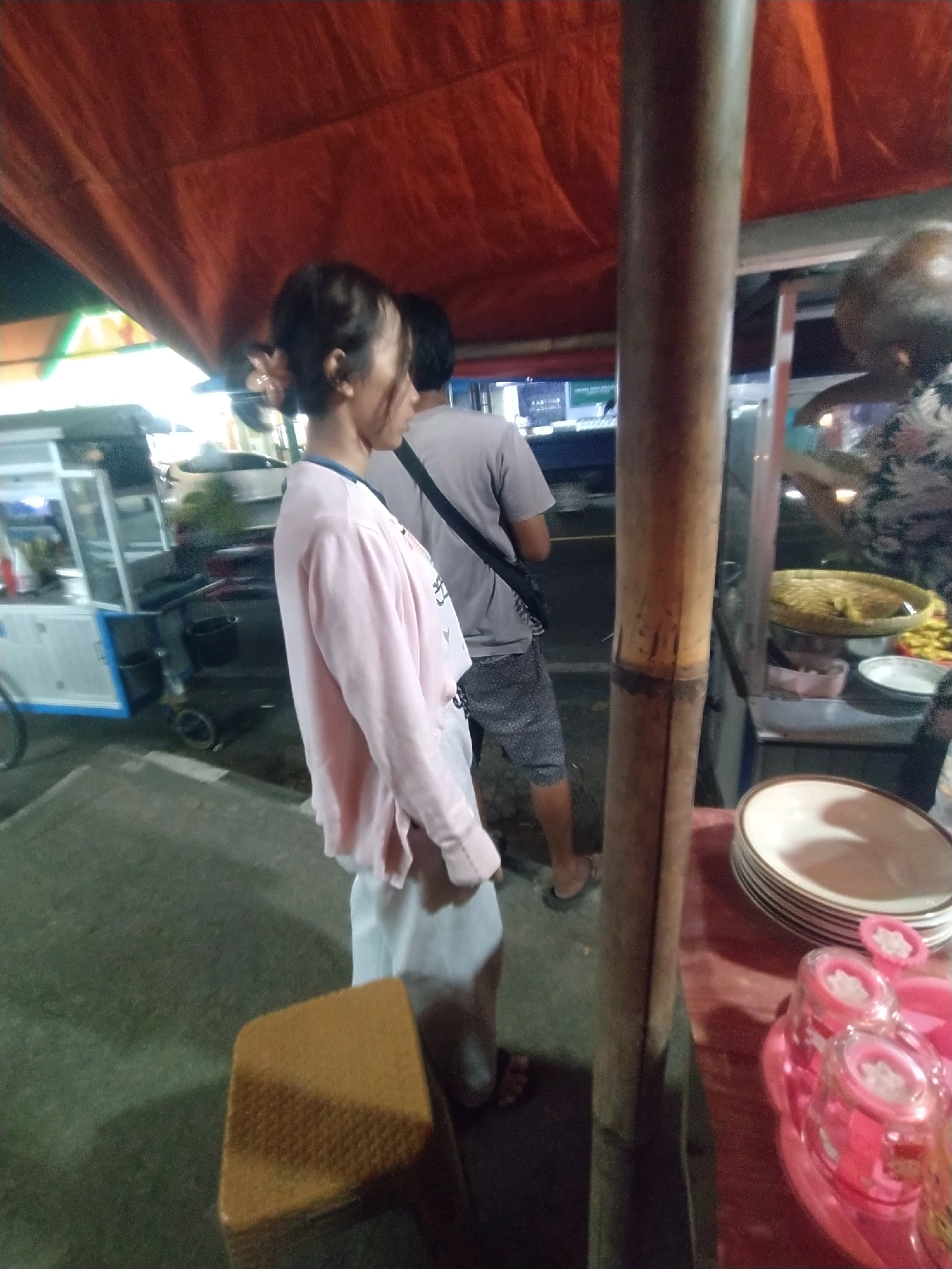 The image depicts an outdoor scene at night in Desa Karangbenda, Indonesia, likely a market or food stall area. A young woman with her hair pulled back in a bun, adorned with a pink flower, stands facing away from the camera, wearing a light pink long-sleeved shirt and white pants. To her right, a man in a grey t-shirt and patterned shorts is also facing away from the camera. They appear to be near a food stall with a red awning overhead and a wooden pole in the foreground. On a table to the right, a stack of plates and some pink condiment containers are visible. The ground is paved and wet, suggesting recent rain or a damp environment. A food cart or stall structure is partially visible on the left side of the image.