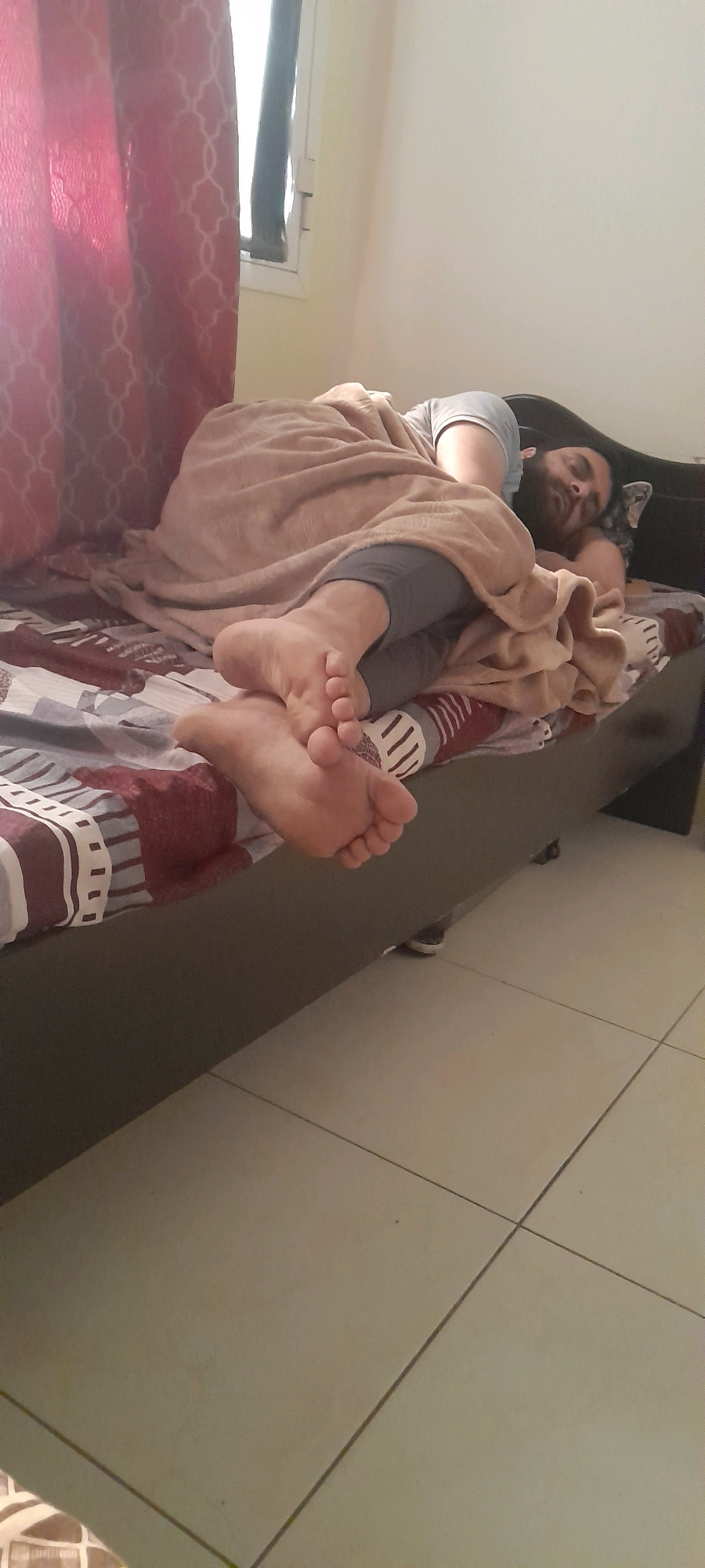 A man with a beard is sleeping on a bed. He is covered by a light brown blanket and wearing grey leggings and a light blue t-shirt. His feet are visible at the end of the bed, with the left foot resting on top of the right. The bed has a patterned duvet with stripes of maroon, white, and grey. A red patterned curtain is visible to the left of the frame, and a window is partially obscured by it. The floor is tiled with square, light-colored tiles. The scene appears to be indoors, possibly in a bedroom, and the lighting suggests it is daytime. The image was taken from a slightly elevated perspective, looking down at the sleeping man and the bed. There is no visible indication of the location being in Doha, Qatar.