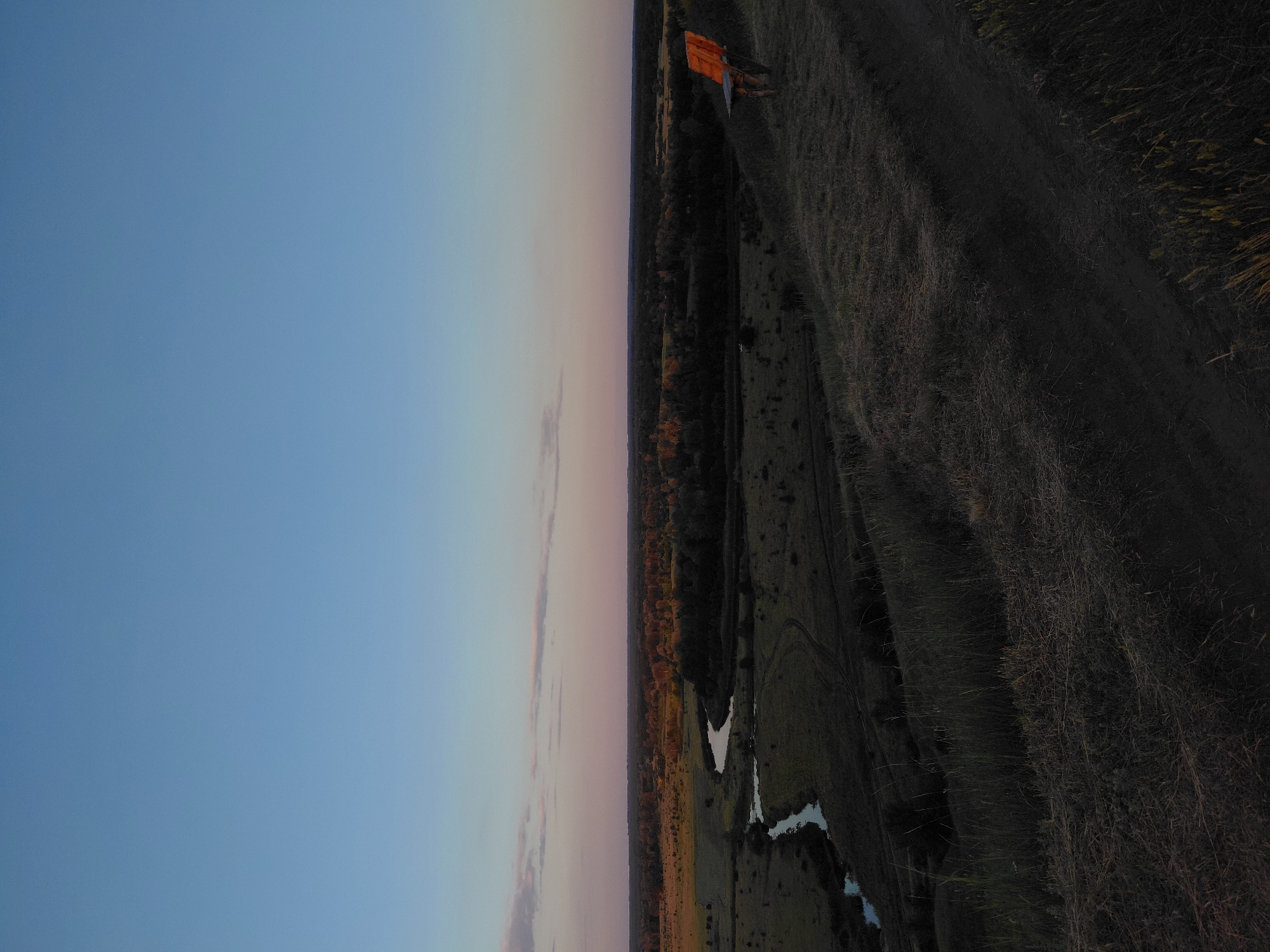 The image depicts a panoramic view of a rural landscape at dusk, likely in or near the village of Ustyvytsia, Ukraine. The sky is a gradient of soft blue to pale pink, with subtle hints of clouds on the horizon. Below the sky, a wide expanse of flat land stretches out, characterized by scattered trees, fields, and a winding river. The foreground features a grassy slope with a dirt path leading down towards the river. On the right side of the slope, a rustic wooden bench is positioned, facing the landscape. The overall scene is serene and quiet, bathed in the fading light of the day.