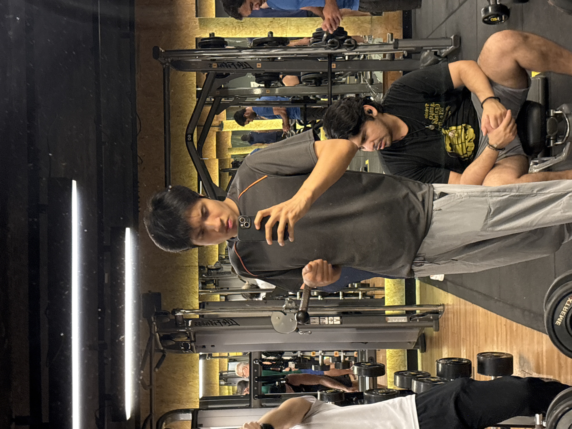 A brightly lit gym, possibly in São Paulo, Brazil, contains several individuals using exercise equipment. In the foreground, a person is taking a selfie with a phone. Adjacent to this person is another individual on a weight machine. In the background, several other people are also engaged in exercise activities. Multiple weight machines and free weights are visible, suggesting a well-equipped fitness facility. The gym has a clean and modern aesthetic.