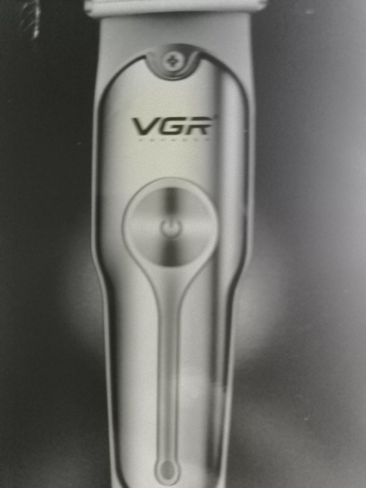 The image shows a close-up, black and white photograph of a silver electric hair trimmer. The trimmer has the brand name "VGR" prominently displayed in black lettering on its body. Below the brand name, there is a circular power button with a power symbol. The trimmer is sleek and metallic, with a rounded top where a screw is visible, and tapers down to a narrower end with a small loop. The background is dark and out of focus, suggesting the trimmer is the sole subject of the photograph. There is no indication of Duttlenheim, France, in the image.
